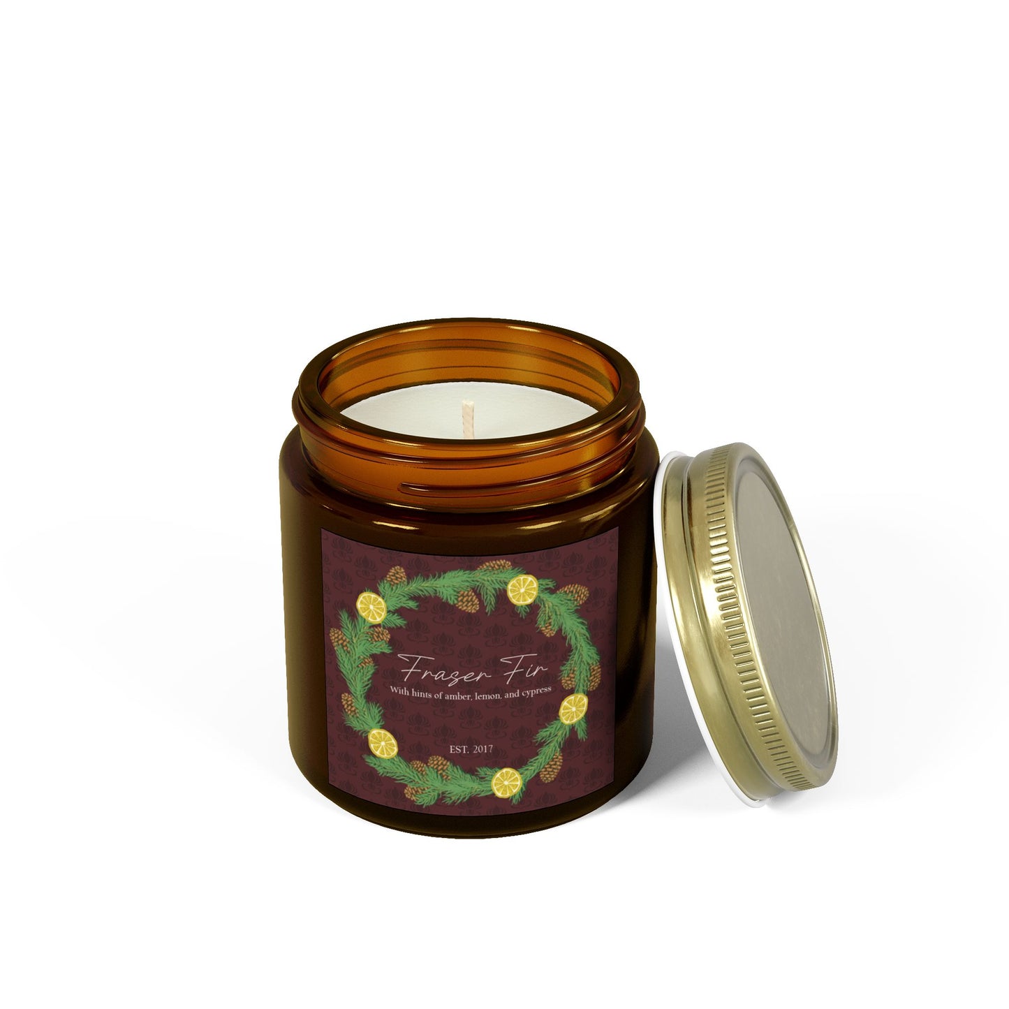 This is the 4oz Amber Fraser Fir Candle from Chris Foster Design. This is a photo from the front open with the tin lid leaning on the right of the glass container. The wick is not lit and the wax is white. The candle is in a glass cylinder container that is tinted amber. The label is brown with a delicate pattern and has a lemon and pinecone wreath surround the name of the candle. The candle is against a white background.
