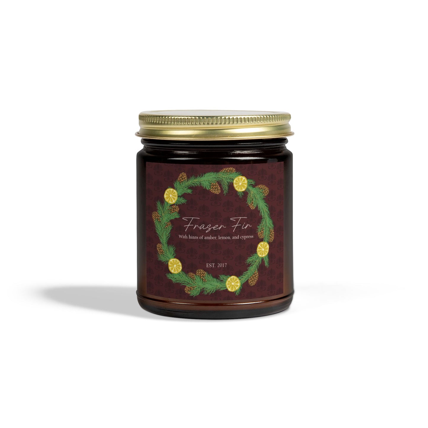 This is the 4oz Amber Fraser Fir Candle from Chris Foster Design. This is a photo from the front with the gold tin lid screwed onto the glass container of the candle. The candle is in a glass cylinder container that is tinted amber. The label is brown with a delicate pattern and has a lemon and pinecone wreath surround the name of the candle. The candle is against a white background.