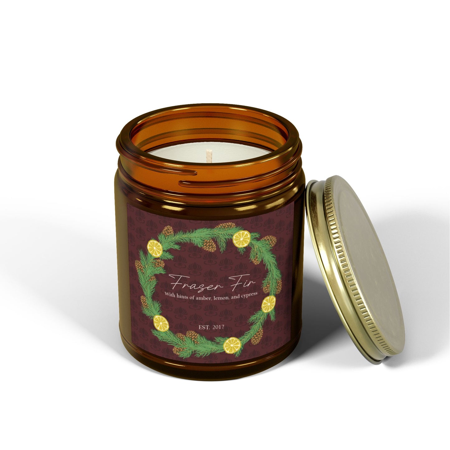 This is the 9oz Amber Fraser Fir Candle from Chris Foster Design. This is a photo from the front open with the tin lid leaning on the right of the glass container. The wick is not lit and the wax is white. The candle is in a glass cylinder container that is tinted amber. The label is brown with a delicate pattern and has a lemon and pinecone wreath surround the name of the candle. The candle is against a white background.