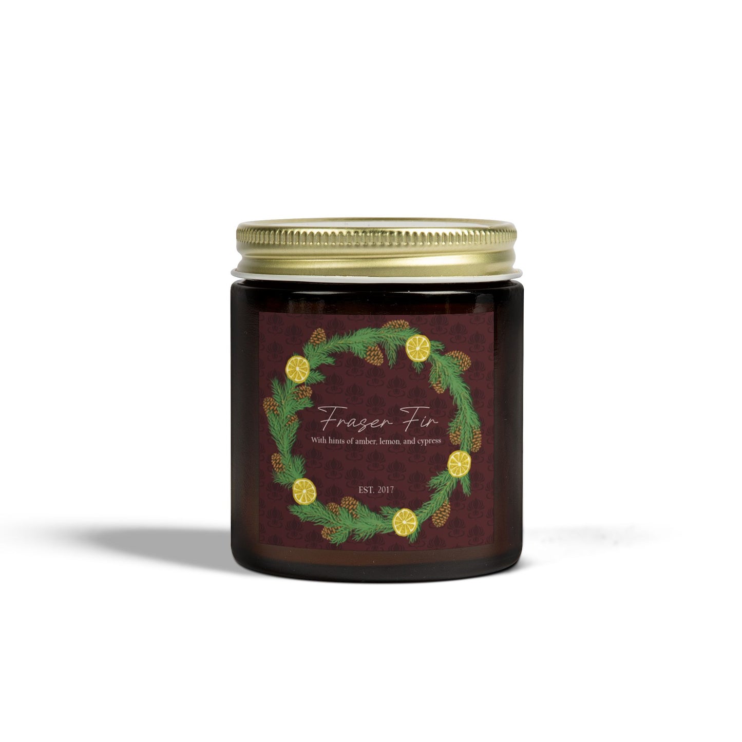 This is the 9oz Amber Fraser Fir Candle from Chris Foster Design. This is a photo from the front with the gold tin lid screwed onto the glass container of the candle. The candle is in a glass cylinder container that is tinted amber. The label is brown with a delicate pattern and has a lemon and pinecone wreath surround the name of the candle. The candle is against a white background.