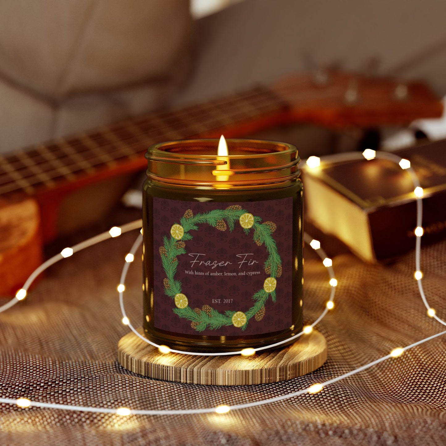 This is the 9oz Amber Fraser Fir Candle from Chris Foster Design. This is a photo from the front open and the wick lit. The candle is in a glass cylinder container that is tinted amber. The label is brown with a delicate pattern and has a lemon and pinecone wreath surround the name of the candle. The candle is on a wood coaster surrounded by little lights. It is on a clothed surface and in the background there is a light wooded ukulele on the left and a brown book on the right and a tan leather sofa.