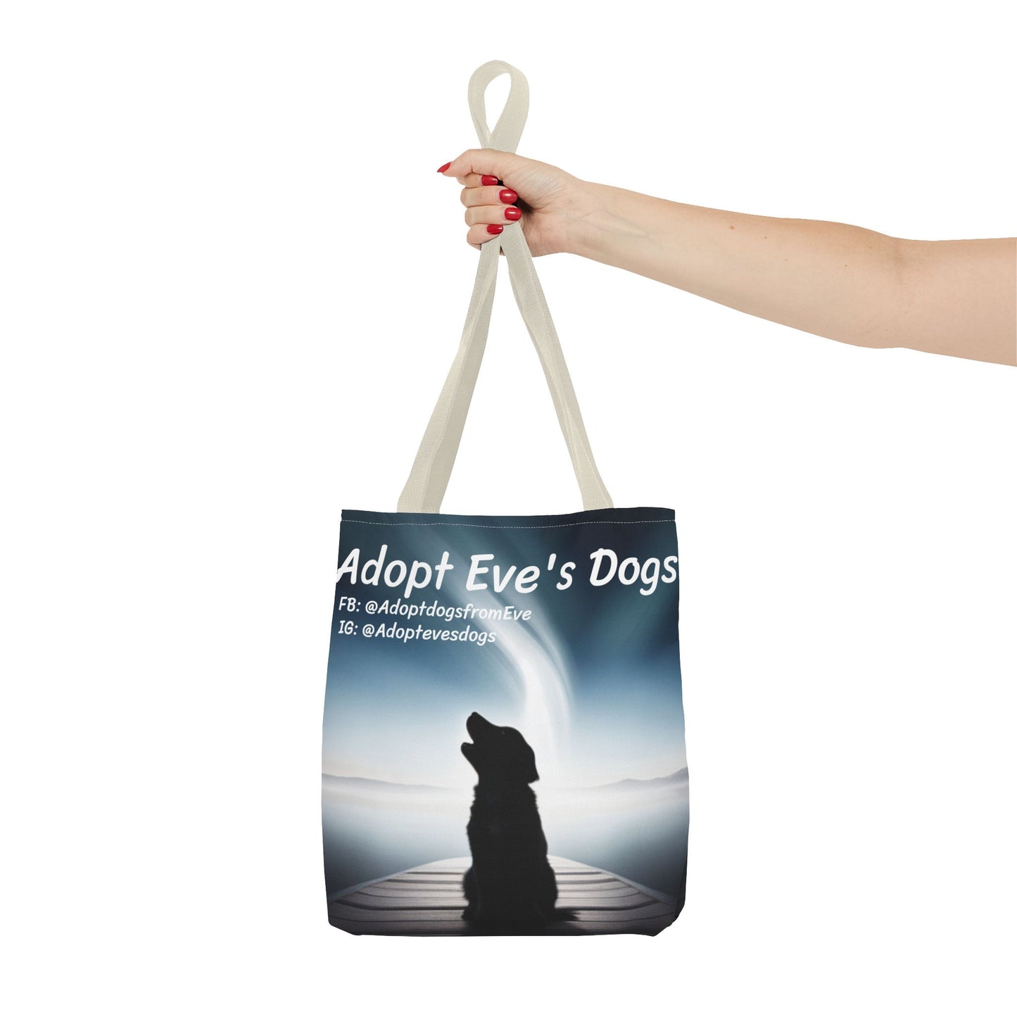This is the Beige 13" x 13" Adopt Eve's Dogs Tote with color matched handles. This is an all around print tote with "Adopt Eve's Dogs" and socials on the top in white. The print is of a shadow of a golden retriever looking to the left on a pier towards water with mountains surrounding it. The image is shown with the upper arm to the hand. The hand holding the handles of tote from the right with red nail polish. The tote is against a white background.