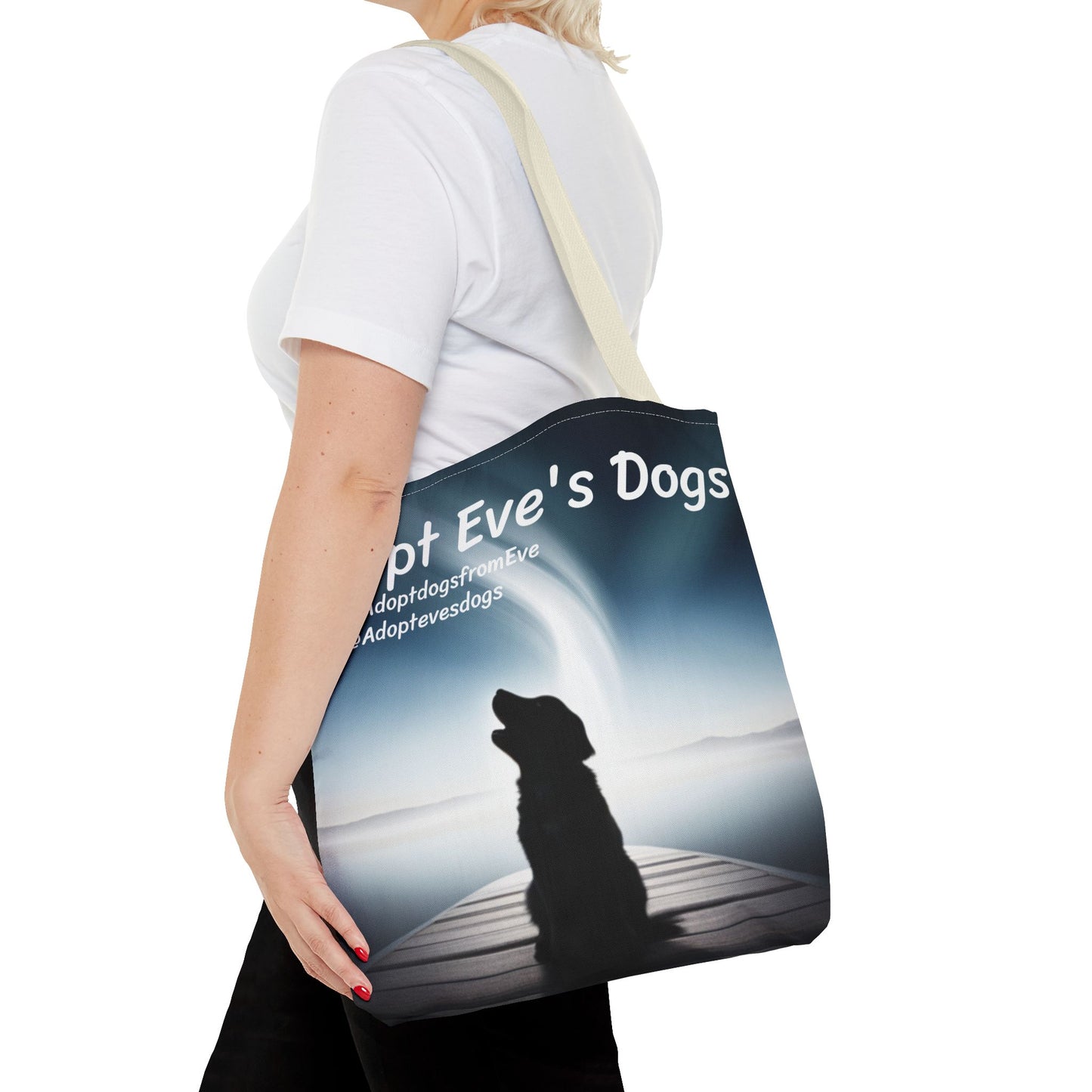 This is the Beige 16" x 16" Adopt Eve's Dogs Tote with color matched handles. This is an all around print tote with "Adopt Eve's Dogs" and socials on the top in white. The print is of a shadow of a golden retriever looking to the left on a pier towards water with mountains surrounding it. There is a woman wearing a white tee, black pants and short blond hair. Her hand is on the tote and has the tote over her shoulder. She is against a white background.