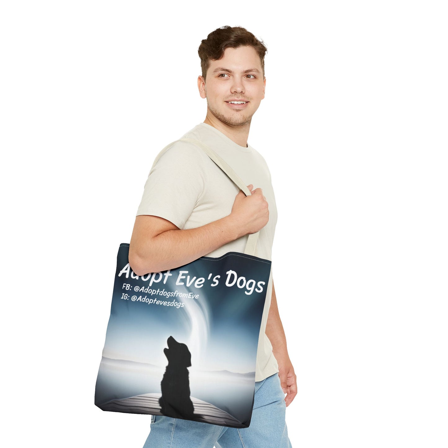 This is the Beige 18" x 18" Adopt Eve's Dogs Tote with color matched handles. This is an all around print tote with "Adopt Eve's Dogs" and socials on the top in white. The print is of a shadow of a golden retriever looking to the left on a pier towards water with mountains surrounding it. There is a smiling brunette man walking to the right, looking to the left, holding the handles of the tote over his shoulder. He is wearing a white tee and light jeans. He is against a white background.