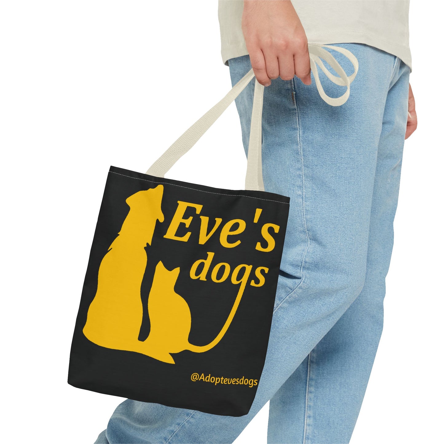 This is the Beige 13" x 13" Adopt Eve's Dog Tote Bag. The handles are beige. The top of the tote is hemmed in yellow. The tote is all over black with the image of a dog, cat, "Eve's Dogs" and "@Adoptevesdogs" in yellow. A man's hand with a beige tee and light- colored jeans is grabbing the handles from the hip down. The tote is against a white background.