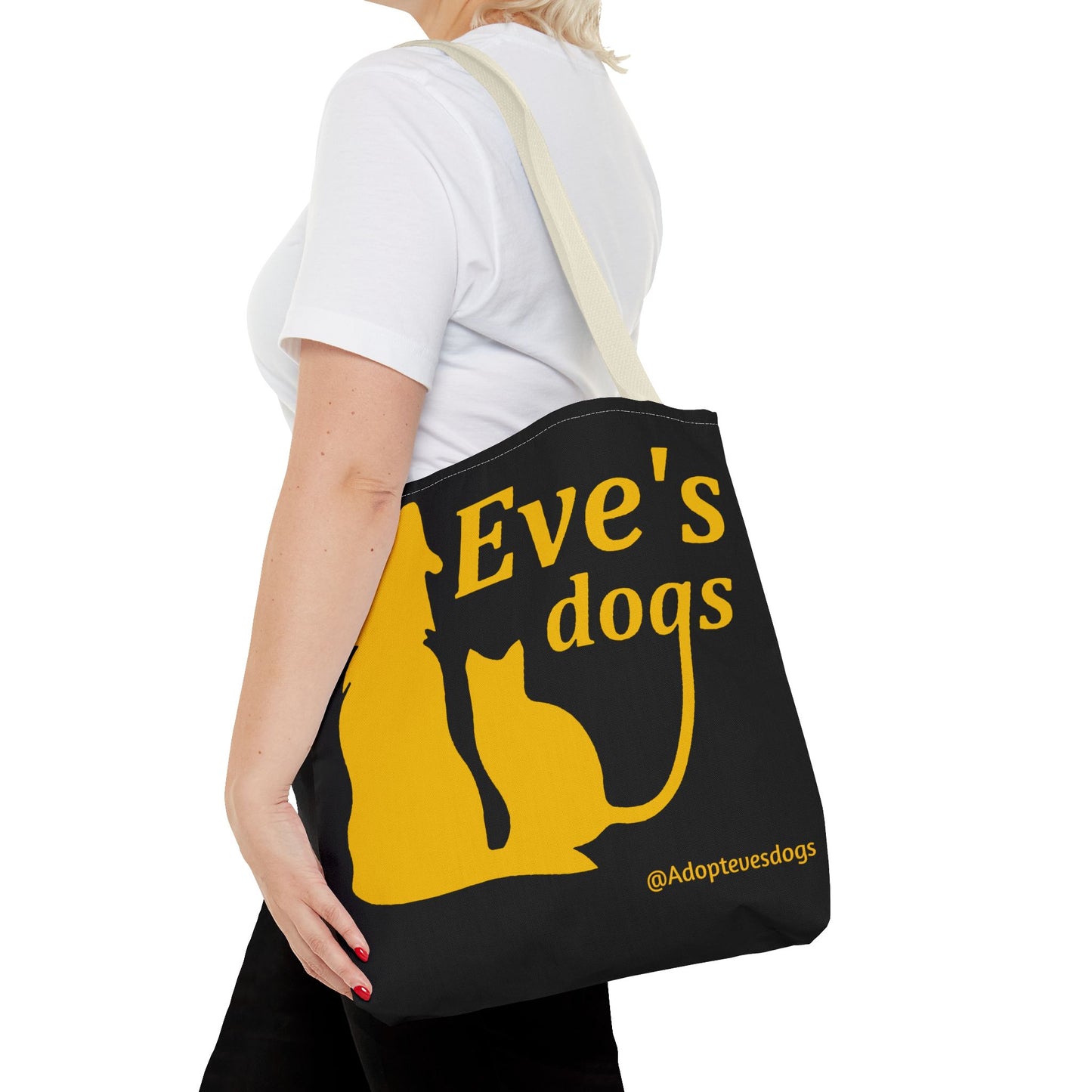 This is the Beige 16" x 16" Adopt Eve's Dog Tote Bag. The handles are beige. The top of the tote is hemmed in yellow. The tote is all over black with the image of a dog, cat, "Eve's Dogs" and "@Adoptevesdogs" in yellow. It is over the shoulder of a short blonde haired woman wearing a white tea and black pants. Her hand with red nails is on the tote and she is facing the left.