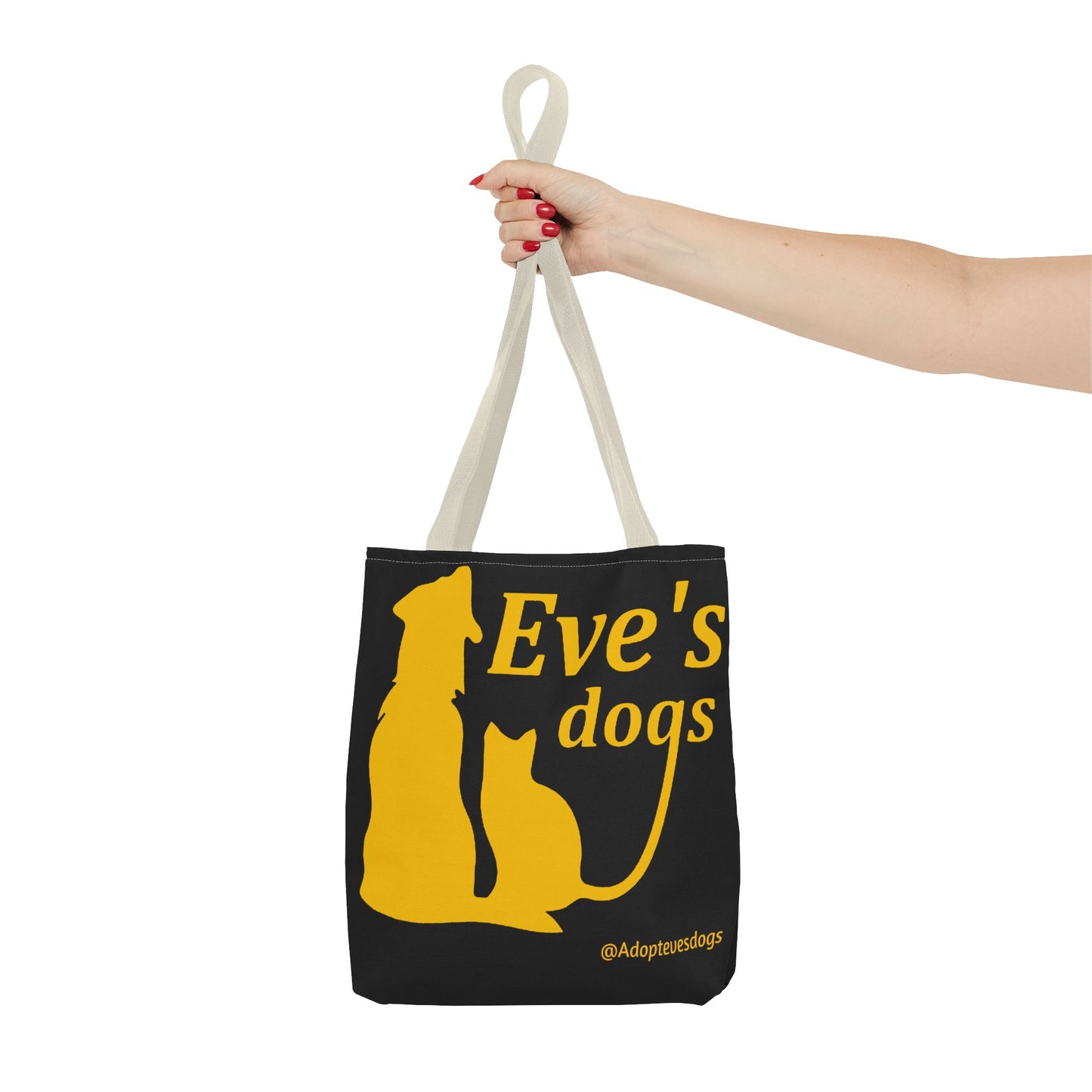 This is the Beige 16" x 16" Adopt Eve's Dog Tote Bag. The handles are beige. The top of the tote is hemmed in yellow. The tote is all over black with the image of a dog, cat, "Eve's Dogs" and "@Adoptevesdogs" in yellow. A woman's hand with red nail polish is grabbing the handles. Her arm is outstretched to the middle. The tote is against a white background.