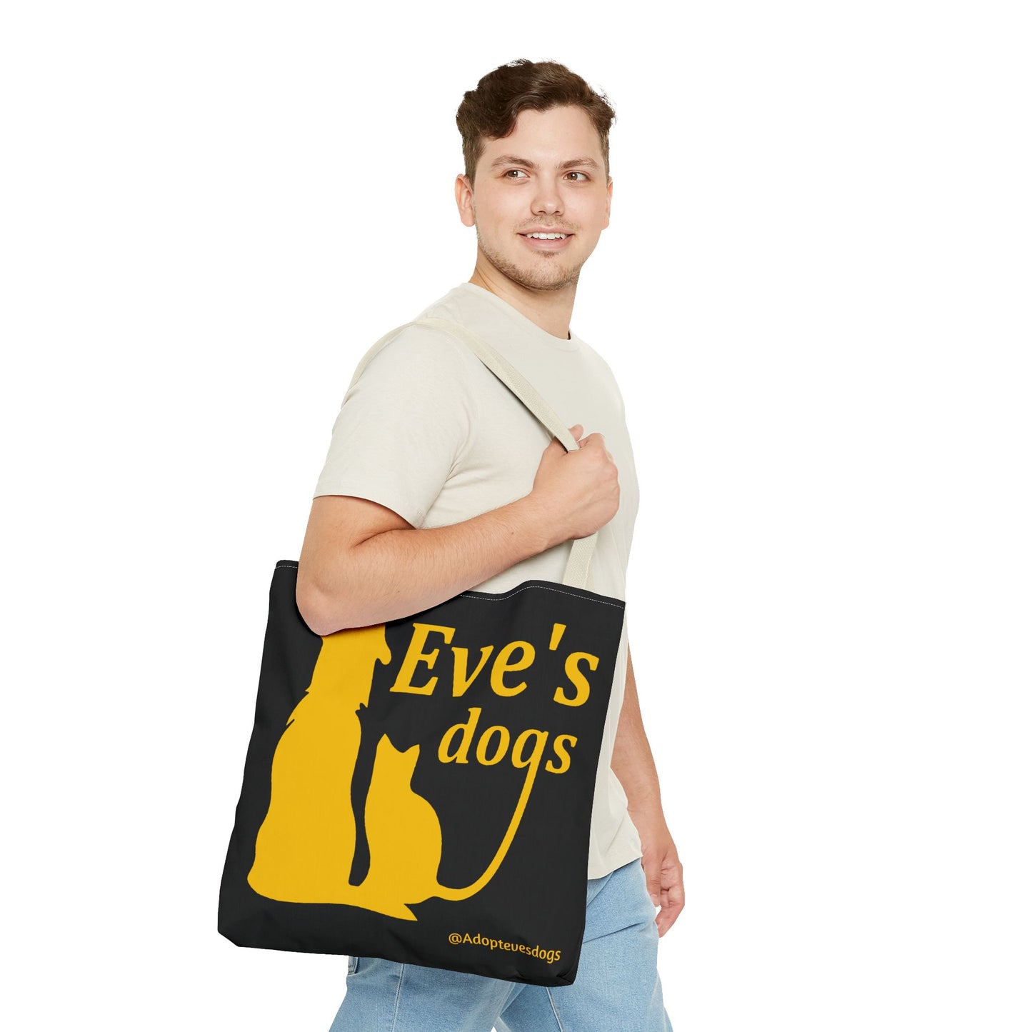 This is the Beige 16" x 16" Adopt Eve's Dog Tote Bag. The handles are beige. The top of the tote is hemmed in yellow. The tote is all over black with the image of a dog, cat, "Eve's Dogs" and "@Adoptevesdogs" in yellow. A man with a smile, short wavy brown hair, beige tee plus jeans with tote around their shoulder and walking to the right against a white background.