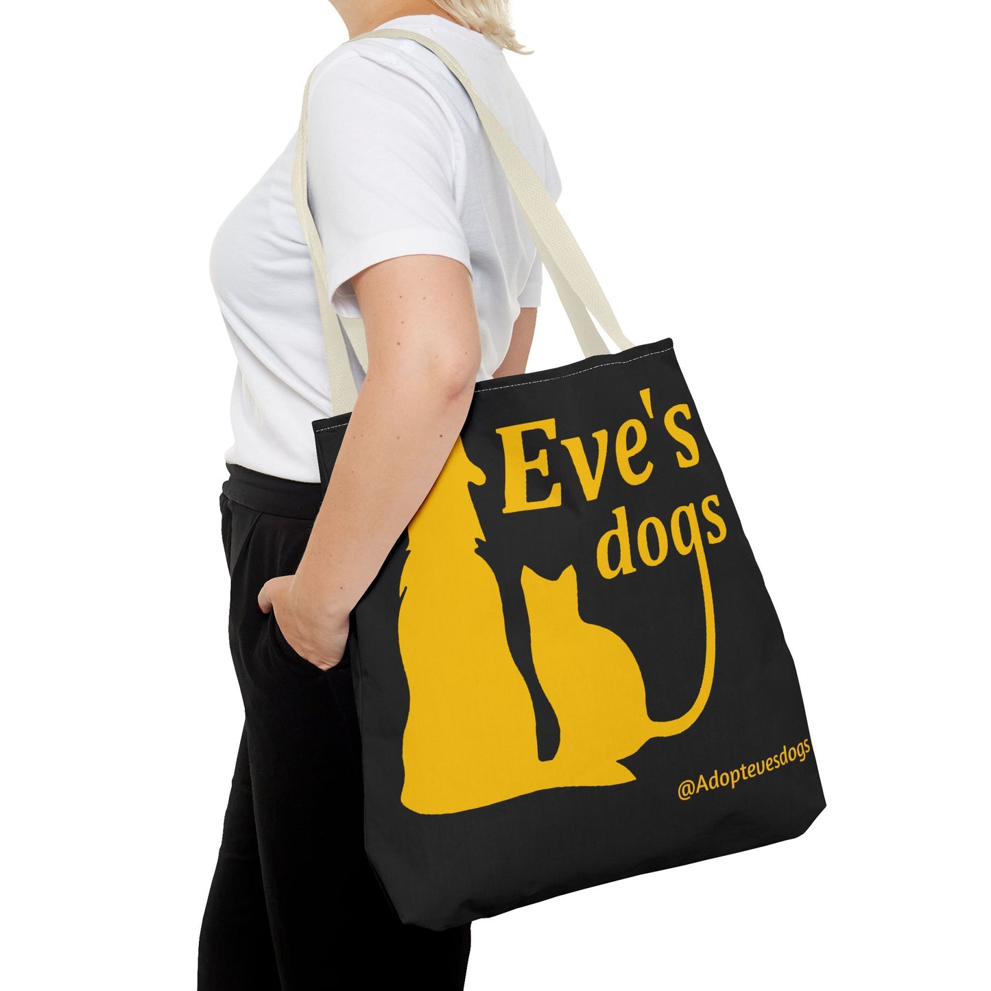 This is the Beige 18" x 18" Adopt Eve's Dog Tote Bag. The handles are beige. The top of the tote is hemmed in yellow. The tote is all over black with the image of a dog, cat, "Eve's Dogs" and "@Adoptevesdogs" in yellow. It is over the shoulder of a short blonde haired woman wearing a white tea and black pants. Her hand is in the pant's pocket and she is facing the left.