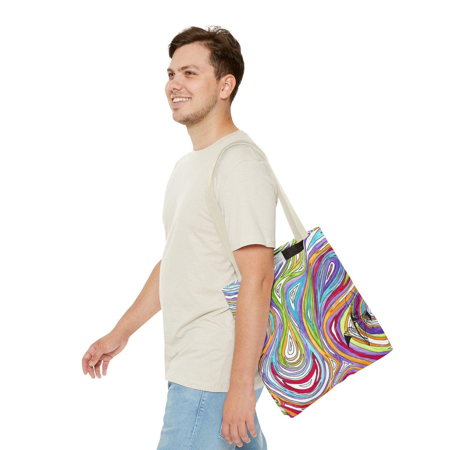 This is the Beige 16" x 16" Oil Spill Tote bag by Storm Garden Studios. A smiling brunette man in white shirt, and blue jeans, side view walking to the left carrying the bag with color matched straps, abstract colors and line work, all over print. The colors are lime green, orange, light blue, dark red, purple, white and black. Storm Garden Studio logo printed in black on bottom right of tote bag. He is against a white background.