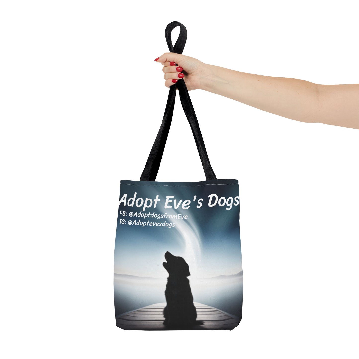 This is the Black 13" x 13" Adopt Eve's Dogs Tote with color matched handles. This is an all around print tote with "Adopt Eve's Dogs" and socials on the top in white. The print is of a shadow of a golden retriever looking to the left on a pier towards water with mountains surrounding it. The image is shown with the upper arm to the hand. The hand holding the handles of tote from the right with red nail polish. The tote is against a white background.