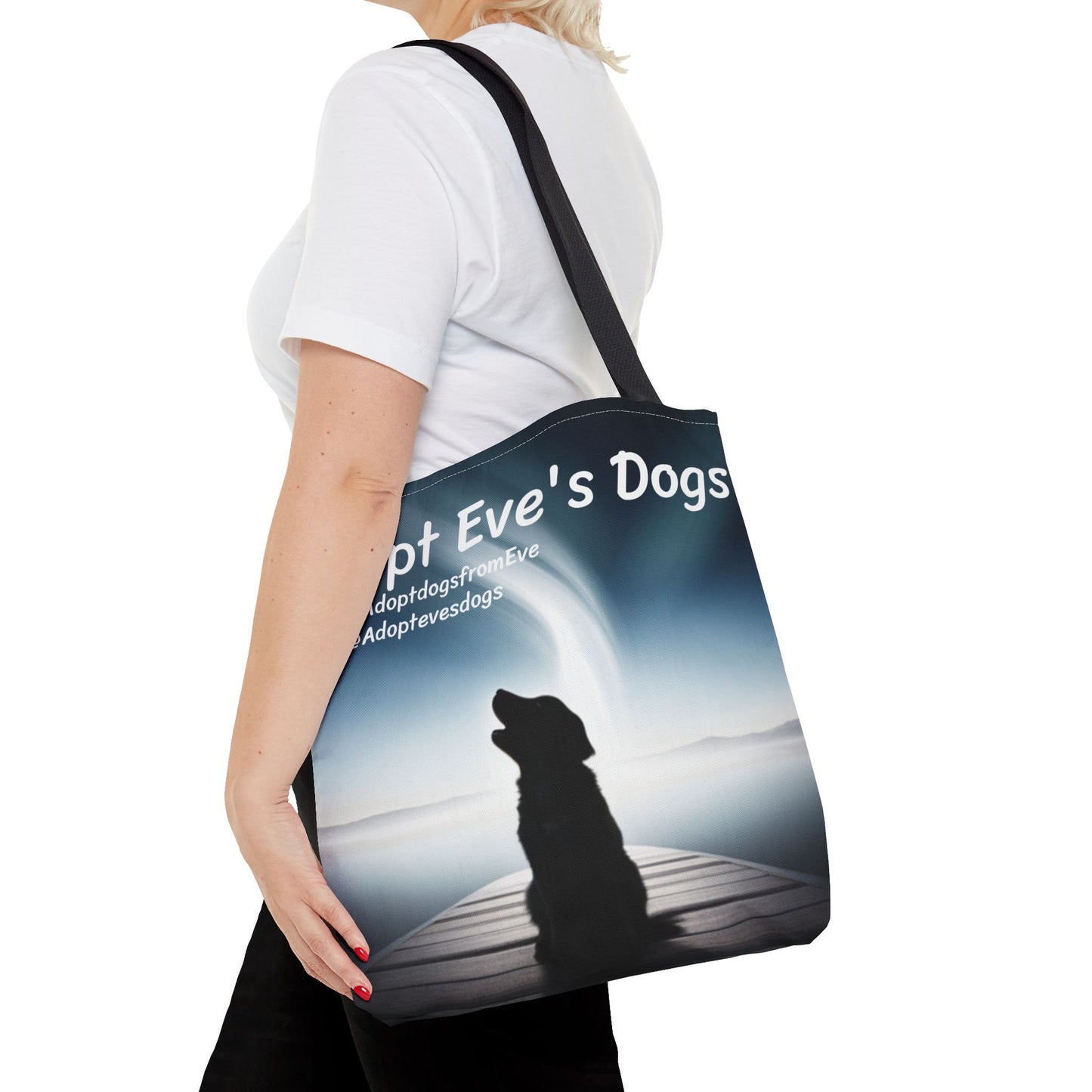 This is the Black 16" x 16" Adopt Eve's Dogs Tote with color matched handles. This is an all around print tote with "Adopt Eve's Dogs" and socials on the top in white. The print is of a shadow of a golden retriever looking to the left on a pier towards water with mountains surrounding it. There is a woman wearing a white tee, black pants and short blond hair. Her hand is on the tote and has the tote over her shoulder. She is against a white background.