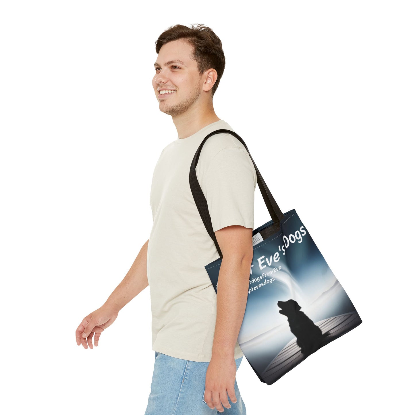 This is the Black 16" x 16" Adopt Eve's Dogs Tote with color matched handles. This is an all around print tote with "Adopt Eve's Dogs" and socials on the top in white. The print is of a shadow of a golden retriever looking to the left on a pier towards water with mountains surrounding it. There is a smiling brunette man walking to the left, looking to the left, with the tote over his shoulder. He is wearing a white tee and light jeans. He is against a white background.