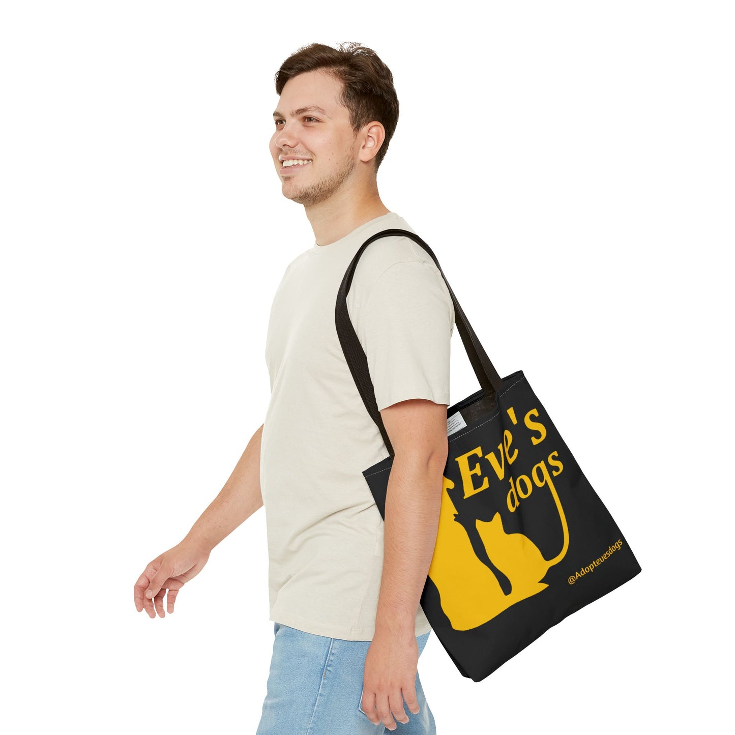 This is the Black 13" x 13" Adopt Eve's Dog Tote Bag. The handles are black. The top of the tote is hemmed in yellow. The tote is all over black with the image of a dog, cat, "Eve's Dogs" and "@Adoptevesdogs" in yellow. A man with a smile, short wavy brown hair, beige tee plus jeans with tote around their shoulder and walking towards the left against a white background.