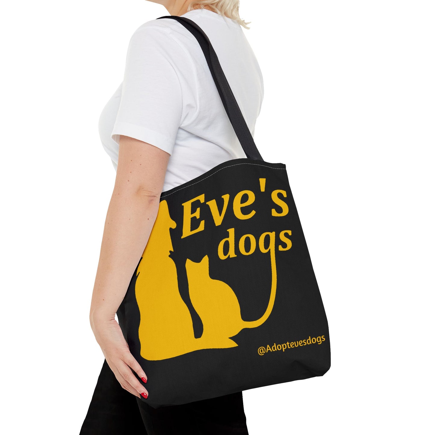 This is the Black 16" x 16" Adopt Eve's Dog Tote Bag. The handles are black. The top of the tote is hemmed in yellow. The tote is all over black with the image of a dog, cat, "Eve's Dogs" and "@Adoptevesdogs" in yellow. It is over the shoulder of a short blonde haired woman wearing a white tea and black pants. Her hand is in the pant's pocket and she is facing the left.