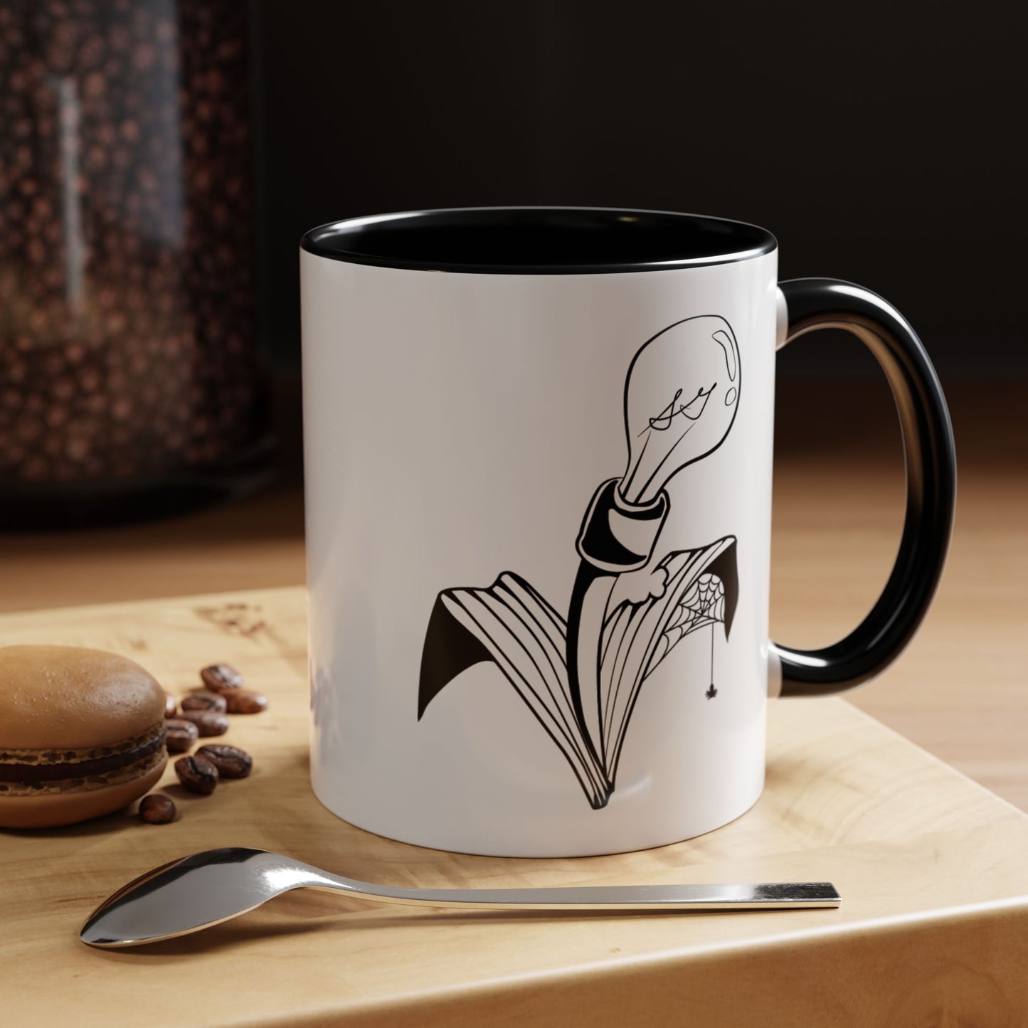 This is the Black 11oz Storm Garden Studio Accent Coffee Mug. This is a mug by Storm Garden Studio which has the logo printed in black and white. The logo is a lightbulb that is screwed onto the leaves of a plant. One leaf is bitten off with a spiderweb. The mug is on top of a light wood block that is on top of a wood desk, a small silver spoon in the front, a chocolate macaroon and some coffee beans to the left. In the background there is a large jar of beans to the left. The wallpaper is black/grey.