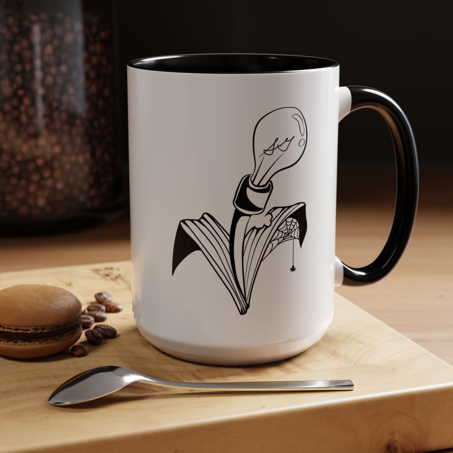 This is the Black 15oz Storm Garden Studio Accent Coffee Mug. This is a mug by Storm Garden Studio which has the logo printed in black and white. The logo is a lightbulb that is screwed onto the leaves of a plant. One leaf is bitten off with a spiderweb. The mug is on top of a light wood block that is on top of a wood desk, a small silver spoon in the front, a chocolate macaroon and some coffee beans to the left. In the background there is a large jar of beans to the left. The wallpaper is black/grey.