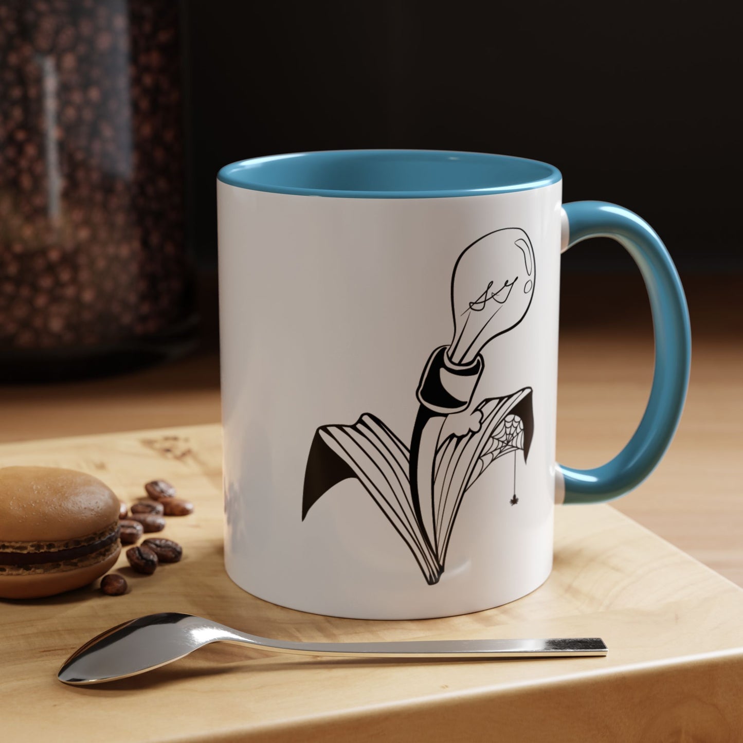 This is the Light Blue 11oz Storm Garden Studio Accent Coffee Mug. This is a mug by Storm Garden Studio which has the logo printed in black and white. The logo is a lightbulb that is screwed onto the leaves of a plant. One leaf is bitten off with a spiderweb. The mug is on top of a light wood block that is on top of a wood desk, a small silver spoon in the front, a chocolate macaroon and some coffee beans to the left. In the background there is a large jar of beans to the left. The wallpaper is black/grey.