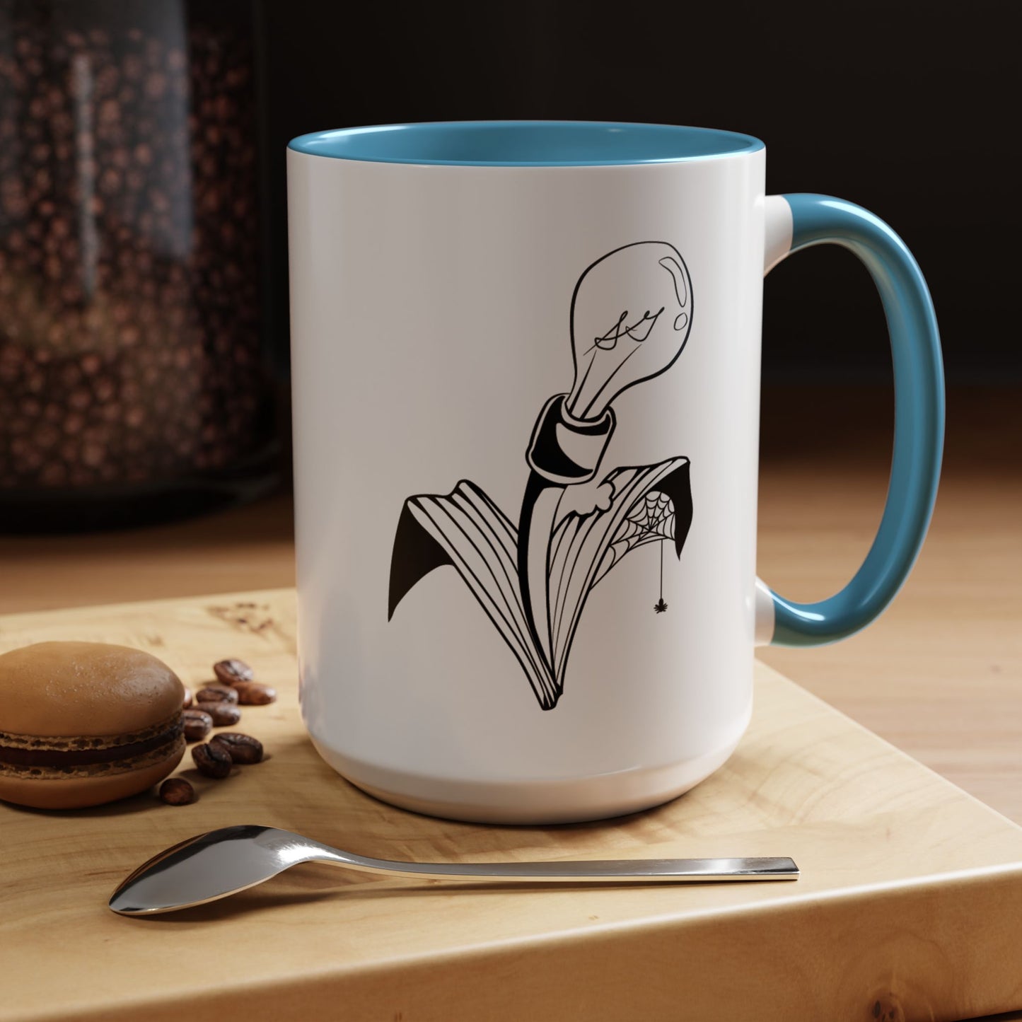 This is the Light Blue 15oz Storm Garden Studio Accent Coffee Mug. This is a mug by Storm Garden Studio which has the logo printed in black and white. The logo is a lightbulb that is screwed onto the leaves of a plant. One leaf is bitten off with a spiderweb. The mug is on top of a light wood block that is on top of a wood desk, a small silver spoon in the front, a chocolate macaroon and some coffee beans to the left. In the background there is a large jar of beans to the left. The wallpaper is black/grey.