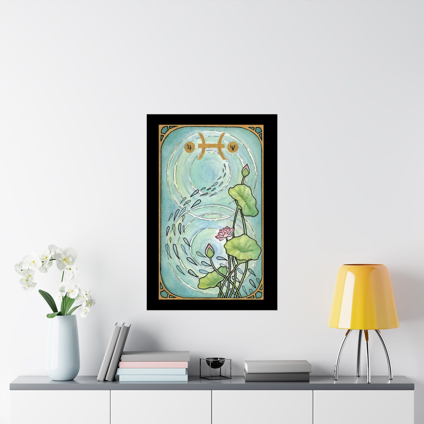This is the Matte 24" x 36" Pisces Poster by Chris Foster Design.The illustration is designed like a tarot card,has a black border,gold frame, a pond,two circular ripples of water,1 bloomed flower,2 buds and 3 leaves combined trailing up on the right with seeds running down the pond,Pisces symbols on the top in gold.Poster is in the middle of a white wall, above gray/white shelf, yellow lamp to the right, blue vase and white flowers to the left, pastel and gray books scattered with one black decoration.