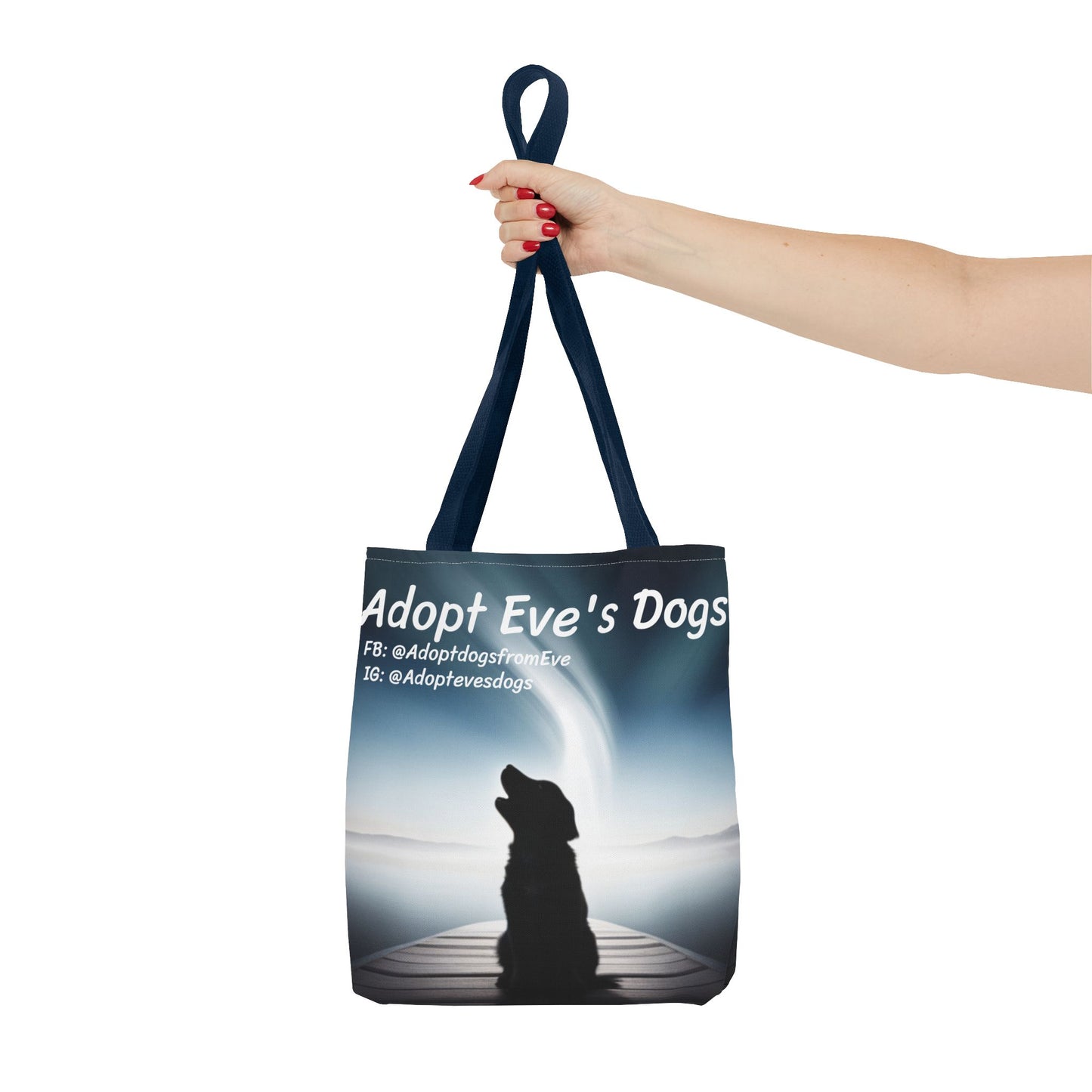 This is the Navy 13" x 13" Adopt Eve's Dogs Tote with color matched handles. This is an all around print tote with "Adopt Eve's Dogs" and socials on the top in white. The print is of a shadow of a golden retriever looking to the left on a pier towards water with mountains surrounding it. The image is shown with the upper arm to the hand. The hand holding the handles of tote from the right with red nail polish. The tote is against a white background.