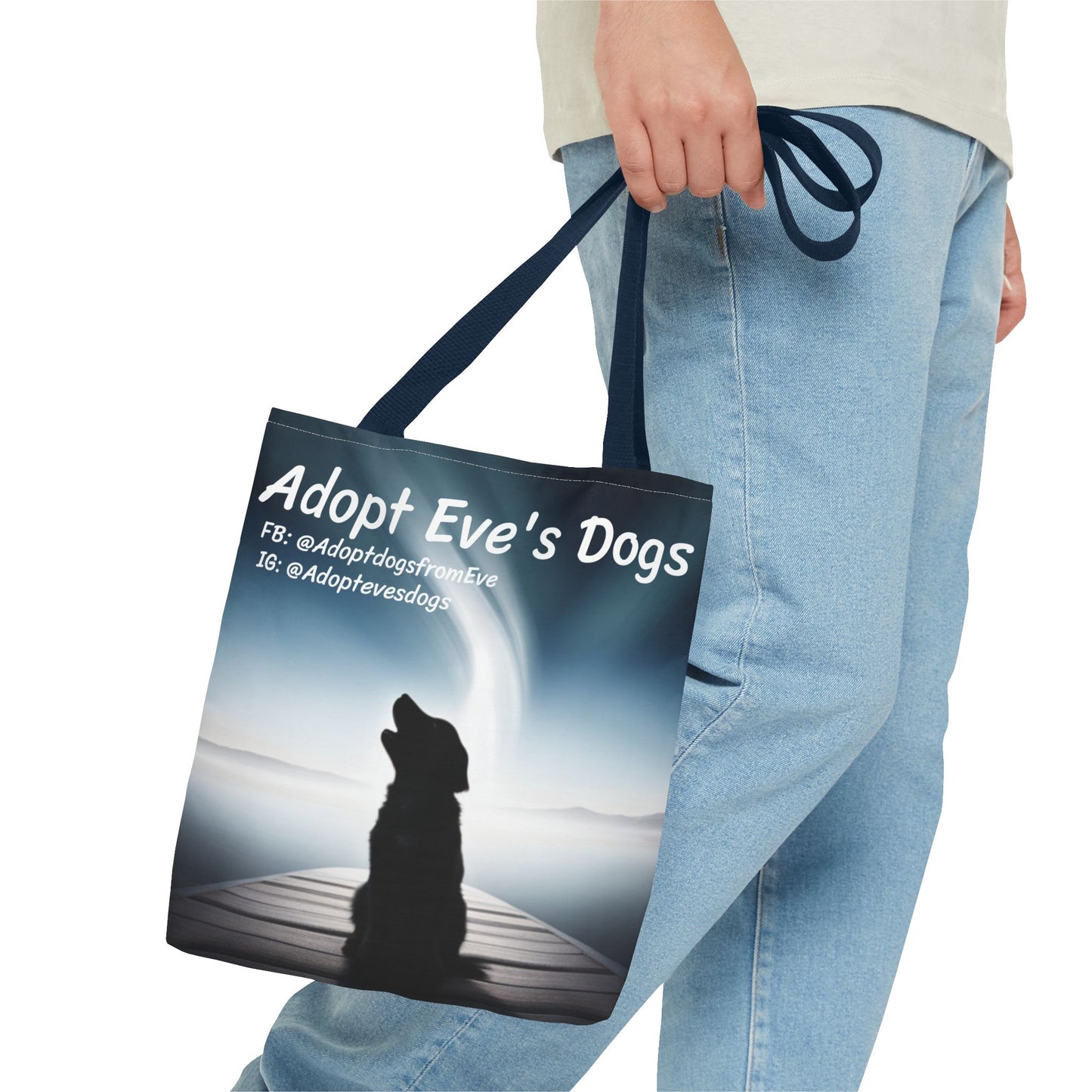 This is the Navy 13" x 13" Adopt Eve's Dogs Tote with color matched handles. This is an all around print tote with "Adopt Eve's Dogs" and socials on the top in white. The print is of a shadow of a golden retriever looking to the left on a pier towards water with mountains surrounding it. This is a close up image from the waist down. A gentleman is holding the tote by the handles wearing a white tee and light jeans walking towards the right.