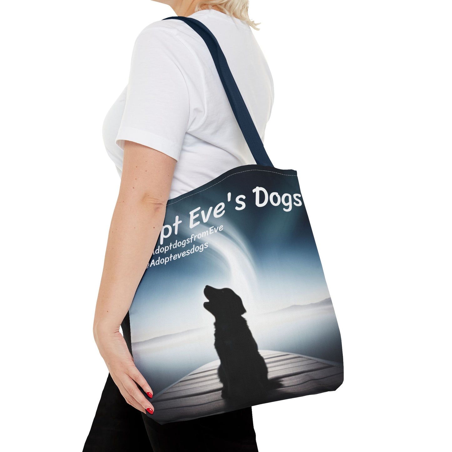 This is the Navy 16" x 16" Adopt Eve's Dogs Tote with color matched handles. This is an all around print tote with "Adopt Eve's Dogs" and socials on the top in white. The print is of a shadow of a golden retriever looking to the left on a pier towards water with mountains surrounding it. There is a woman wearing a white tee, black pants and short blond hair. Her hand is on the tote and has the tote over her shoulder. She is against a white background.