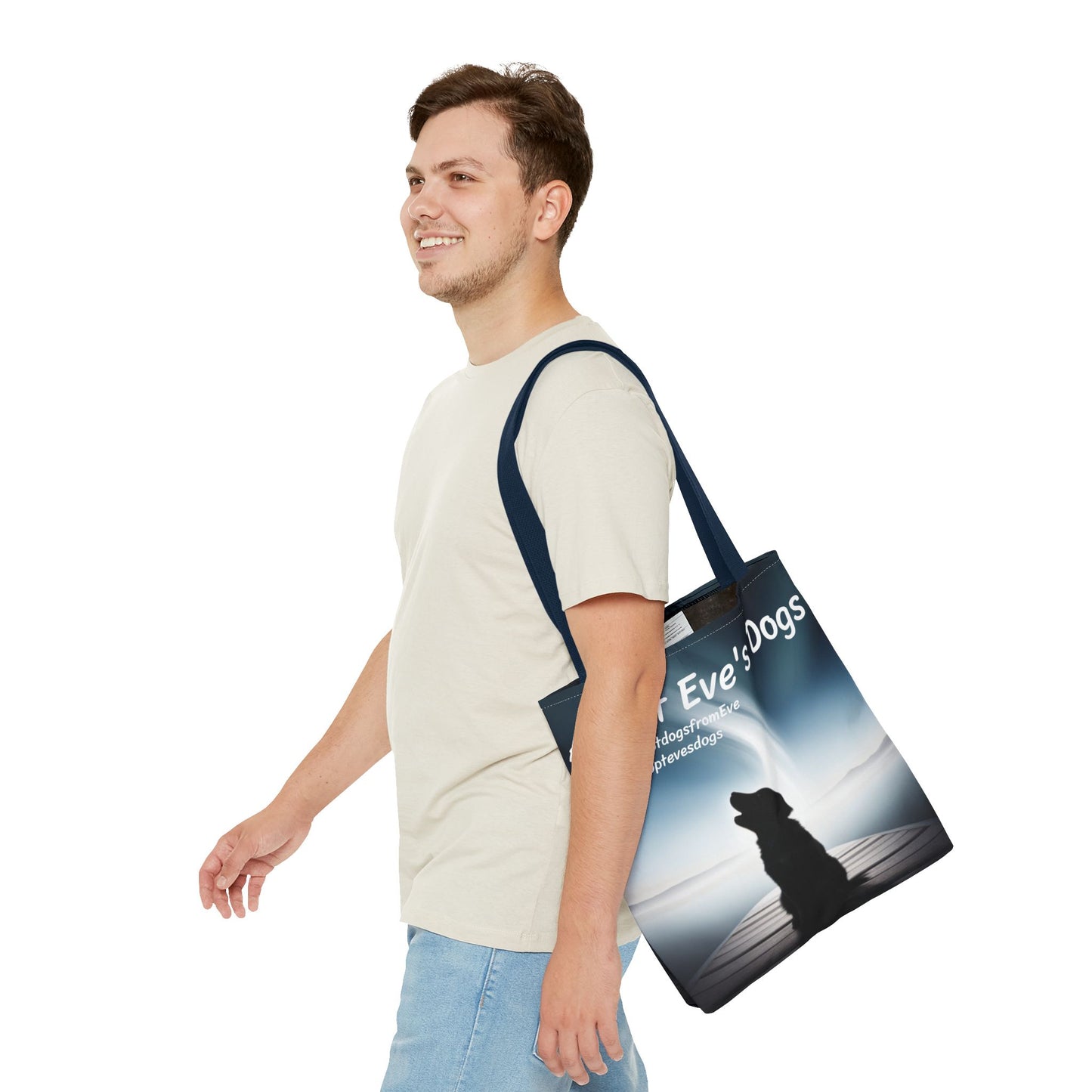 This is the Navy 16" x 16" Adopt Eve's Dogs Tote with color matched handles. This is an all around print tote with "Adopt Eve's Dogs" and socials on the top in white. The print is of a shadow of a golden retriever looking to the left on a pier towards water with mountains surrounding it. There is a smiling brunette man walking to the left, looking to the left, with the tote over his shoulder. He is wearing a white tee and light jeans. He is against a white background.