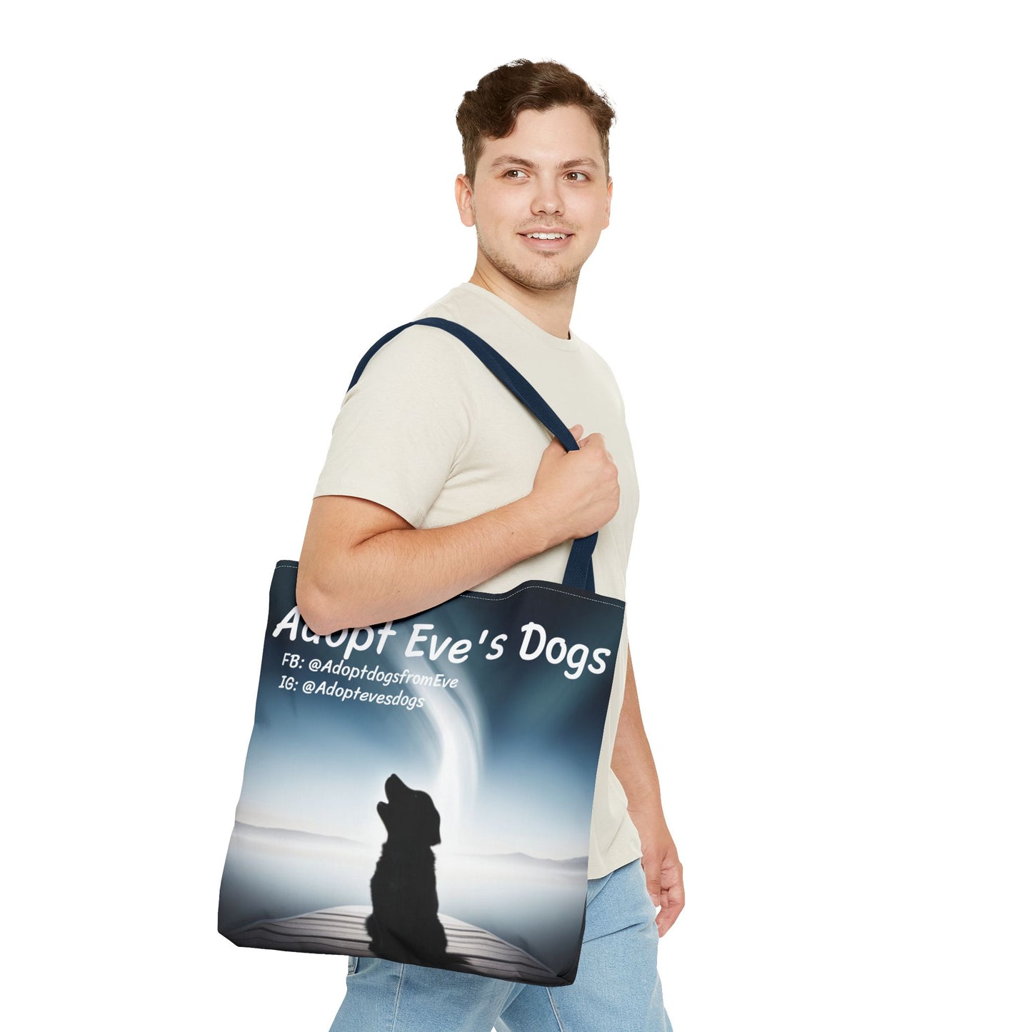 This is the Navy 18" x 18" Adopt Eve's Dogs Tote with color matched handles. This is an all around print tote with "Adopt Eve's Dogs" and socials on the top in white. The print is of a shadow of a golden retriever looking to the left on a pier towards water with mountains surrounding it. There is a smiling brunette man walking to the right, looking to the left, holding the handles of the tote over his shoulder. He is wearing a white tee and light jeans. He is against a white background.