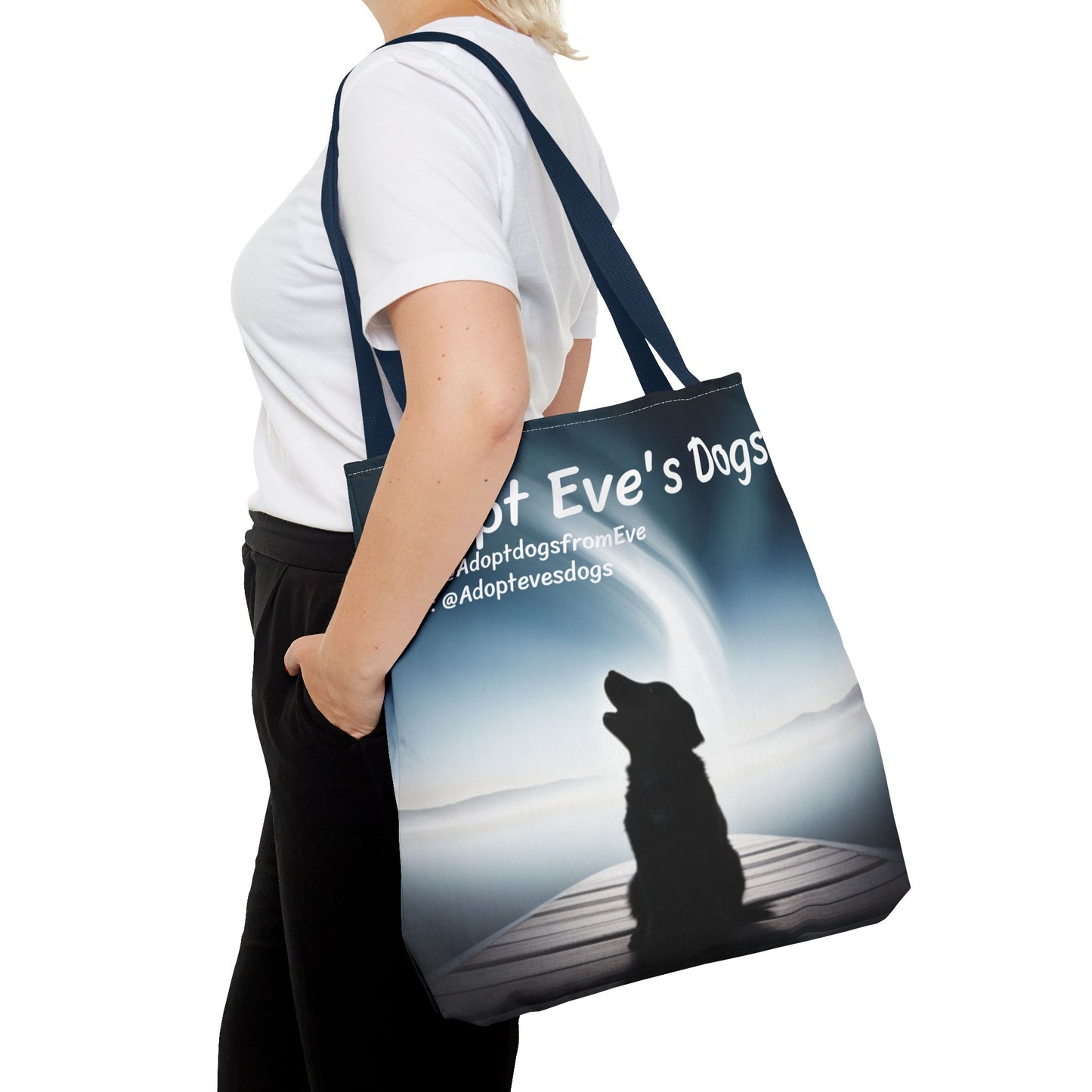 This is the Navy 18" x 18" Adopt Eve's Dogs Tote with color matched handles. This is an all around print tote with "Adopt Eve's Dogs" and socials on the top in white. The print is of a shadow of a golden retriever looking to the left on a pier towards water with mountains surrounding it. There is a woman wearing a white tee, black pants and short blond hair. Her hand is in the pants pocket and has the tote over her shoulder. She is against a white background.