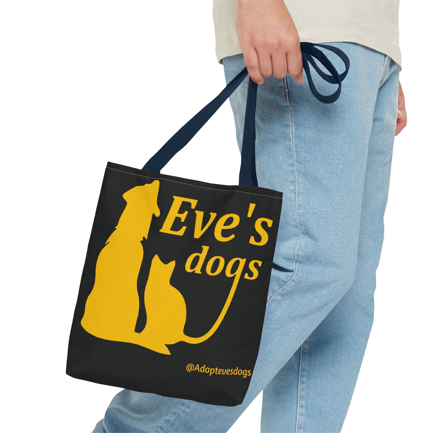 This is the Navy 13" x 13" Adopt Eve's Dog Tote Bag. The handles are navy. The top of the tote is hemmed in yellow. The tote is all over black with the image of a dog, cat, "Eve's Dogs" and "@Adoptevesdogs" in yellow. A man's hand with a beige tee and light- colored jeans is grabbing the handles from the hip down. The tote is against a white background.