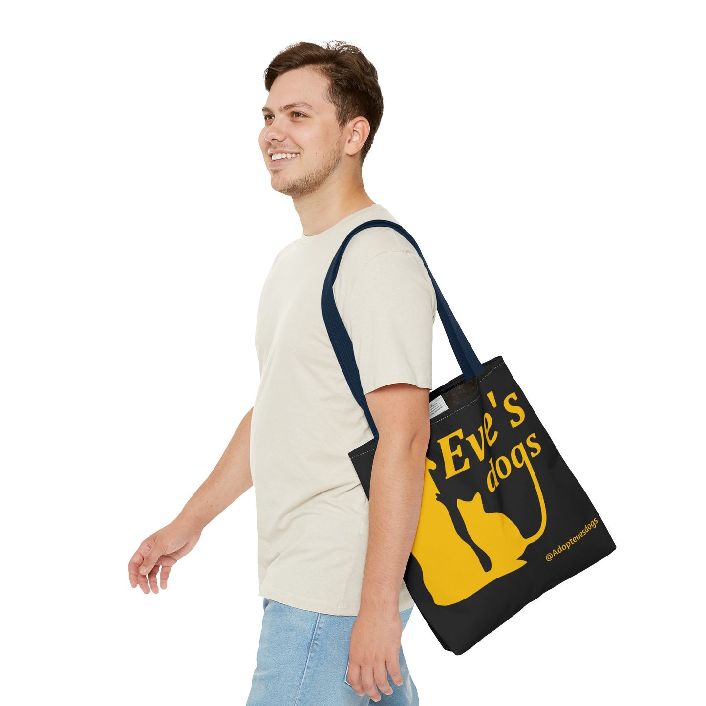 This is the Navy 13" x 13" Adopt Eve's Dog Tote Bag. The handles are navy. The top of the tote is hemmed in yellow. The tote is all over black with the image of a dog, cat, "Eve's Dogs" and "@Adoptevesdogs" in yellow. A man with a smile, short wavy brown hair, beige tee plus jeans with tote around their shoulder and walking towards the left against a white background.