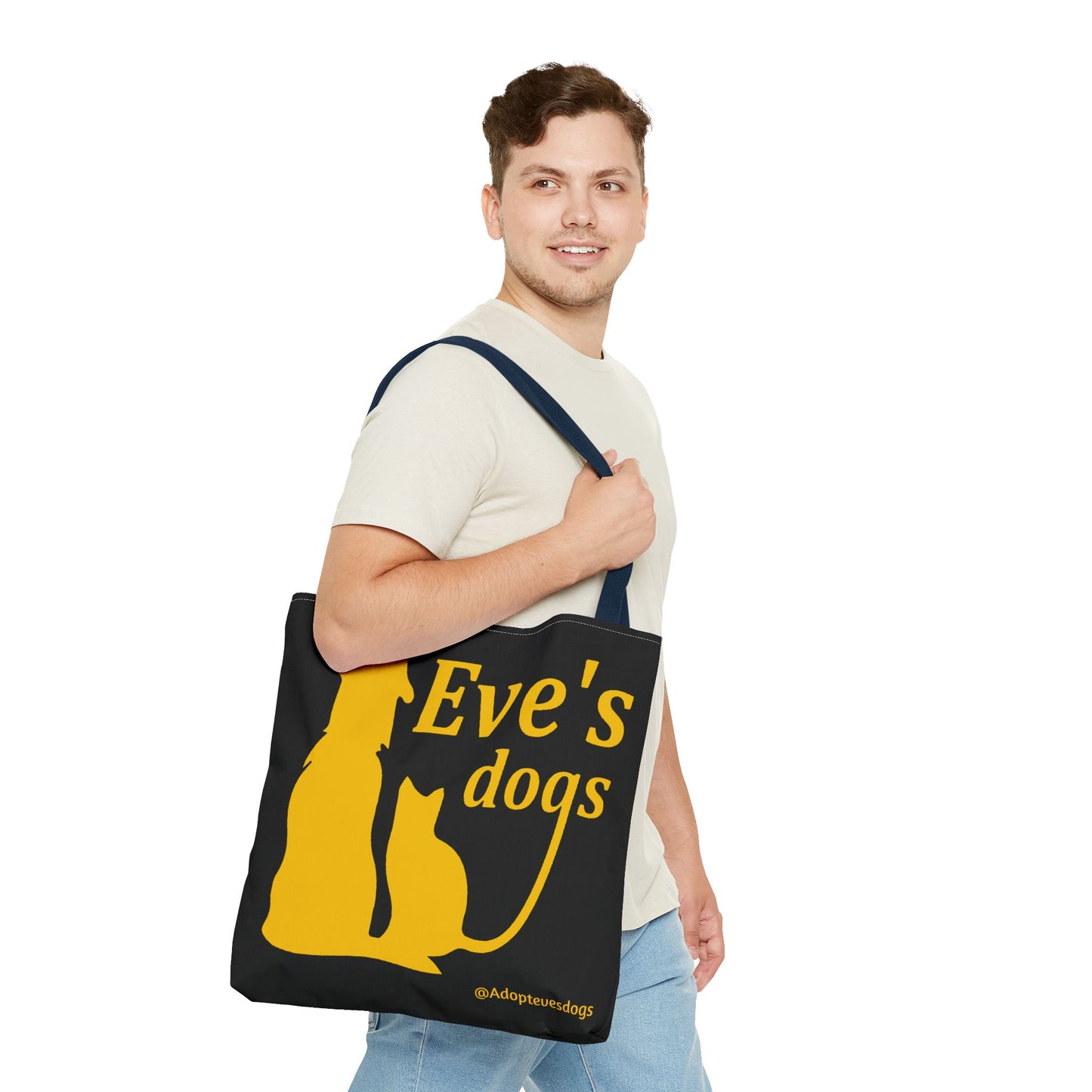 This is the Navy 16" x 16" Adopt Eve's Dog Tote Bag. The handles are navy. The top of the tote is hemmed in yellow. The tote is all over black with the image of a dog, cat, "Eve's Dogs" and "@Adoptevesdogs" in yellow. A man with a smile, short wavy brown hair, beige tee plus jeans with tote around their shoulder and walking to the right against a white background.