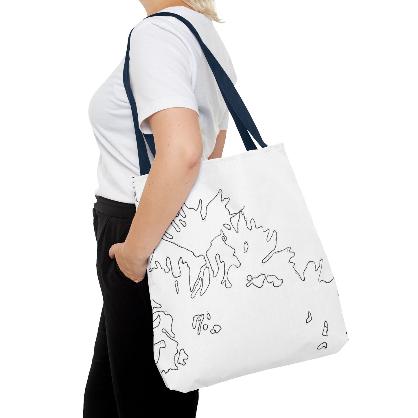This is the Navy 18" x 18" Flowers Tote Bag by Storm Garden Studio. The tote has color matched straps, abstract line work which is outlines of flowers, all over print. The colors are white and black. A blond haired lady is walking towards the left wearing a white tee, black pants, hand in pant's pocket and image is from the shoulders down. She is against a white background.