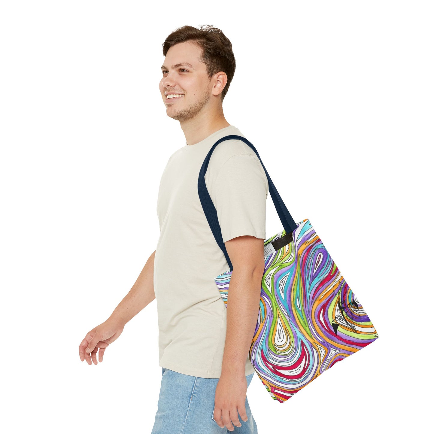 This is the Navy 16" x 16" Oil Spill Tote bag by Storm Garden Studios. A smiling brunette man in white shirt, and blue jeans, side view walking to the left carrying the bag with color matched straps, abstract colors and line work, all over print. The colors are lime green, orange, light blue, dark red, purple, white and black. Storm Garden Studio logo printed in black on bottom right of tote bag. He is against a white background.