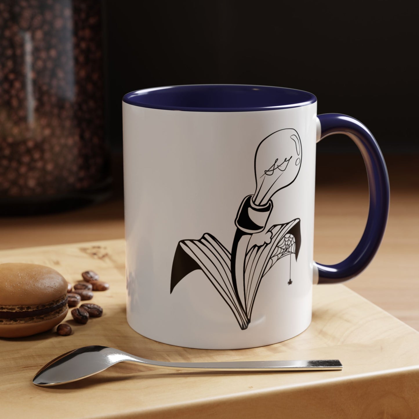 This is the Navy 11oz Storm Garden Studio Accent Coffee Mug. This is a mug by Storm Garden Studio which has the logo printed in black and white. The logo is a lightbulb that is screwed onto the leaves of a plant. One leaf is bitten off with a spiderweb. The mug is on top of a light wood block that is on top of a wood desk, a small silver spoon in the front, a chocolate macaroon and some coffee beans to the left. In the background there is a large jar of beans to the left. The wallpaper is black/grey.