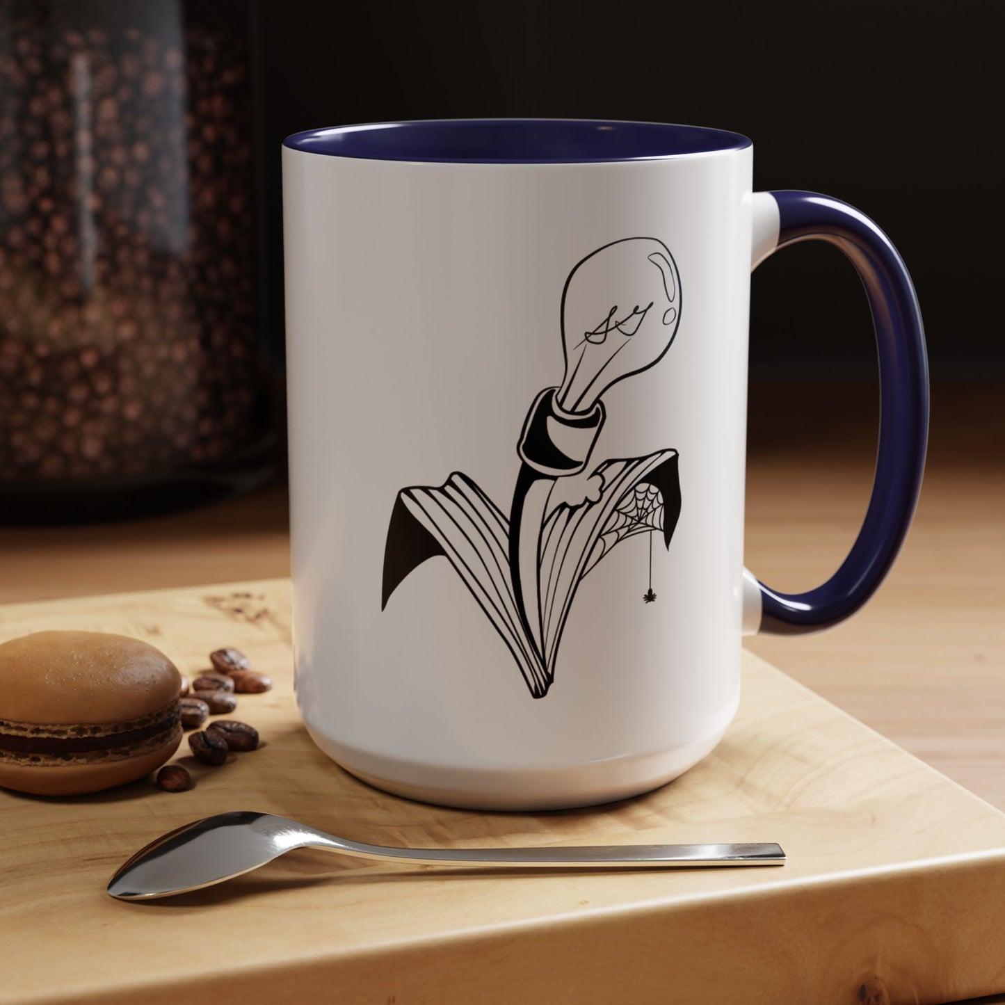 This is the Navy 15oz Storm Garden Studio Accent Coffee Mug. This is a mug by Storm Garden Studio which has the logo printed in black and white. The logo is a lightbulb that is screwed onto the leaves of a plant. One leaf is bitten off with a spiderweb. The mug is on top of a light wood block that is on top of a wood desk, a small silver spoon in the front, a chocolate macaroon and some coffee beans to the left. In the background there is a large jar of beans to the left. The wallpaper is black/grey.