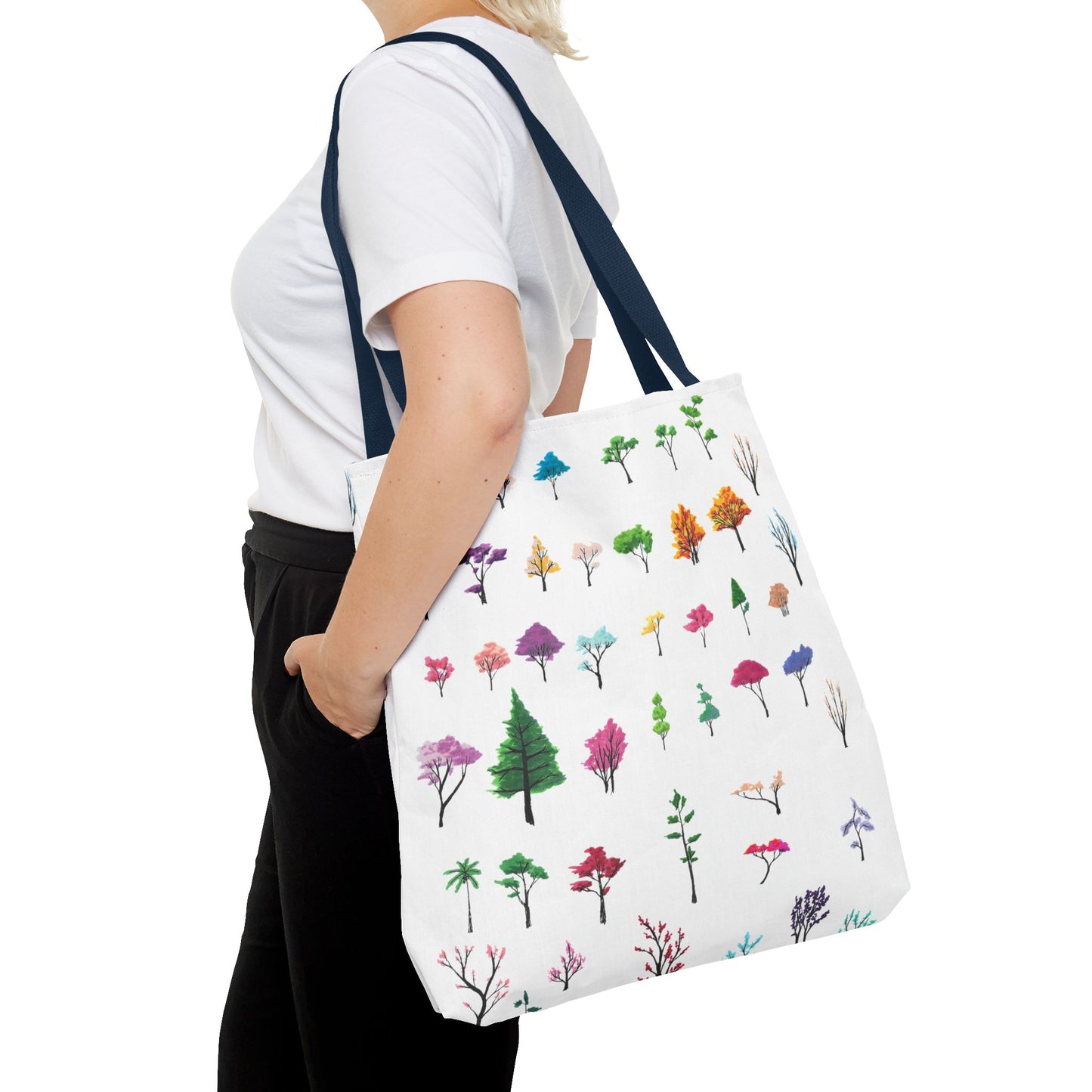 This is the Navy 18" x 18" Study of Trees Tote with color matched handles by Lee Hansheng Studios. There are a variety of tree illustrations from blooming fruit trees to deciduous and evergreens. The trees are colored pinks, purples, greens, yellows, blues and oranges on a white background. The tote is over the shoulder of a short blonde haired woman who has a white tee and black pants. Her hand is in the pocket of the pants and image only shows from the neck to upper thigh.