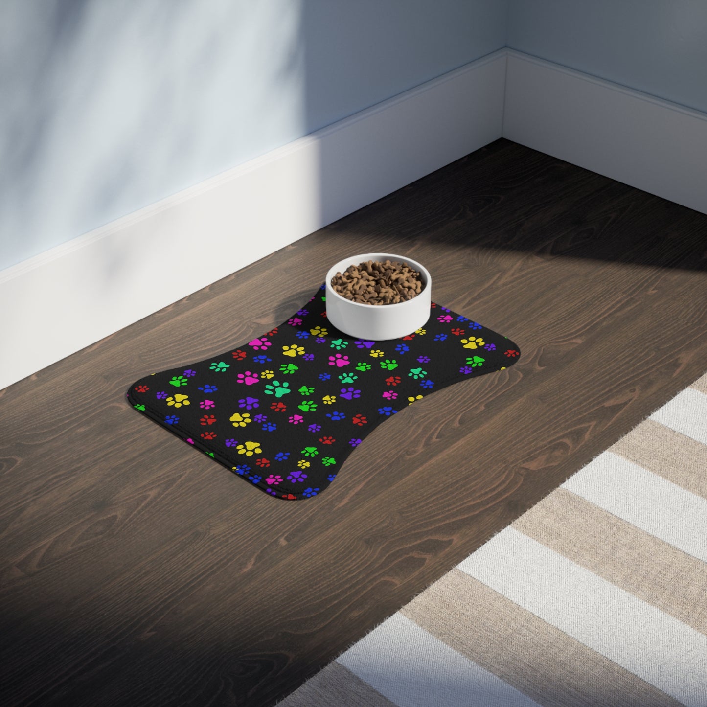 This is the 19" x 14" Bone Shape Paws on Paws Pet Feeding Mat. This shows the bone shape with different sized color paws on the design. The colors are yellow, green red, blue and purple on a black background. There is a white ceramic bowl of food on top of the right corner. The mat is placed on a hard wood floor and right below is a white and tan striped rug. The wall is a light blue and the baseboard is white.