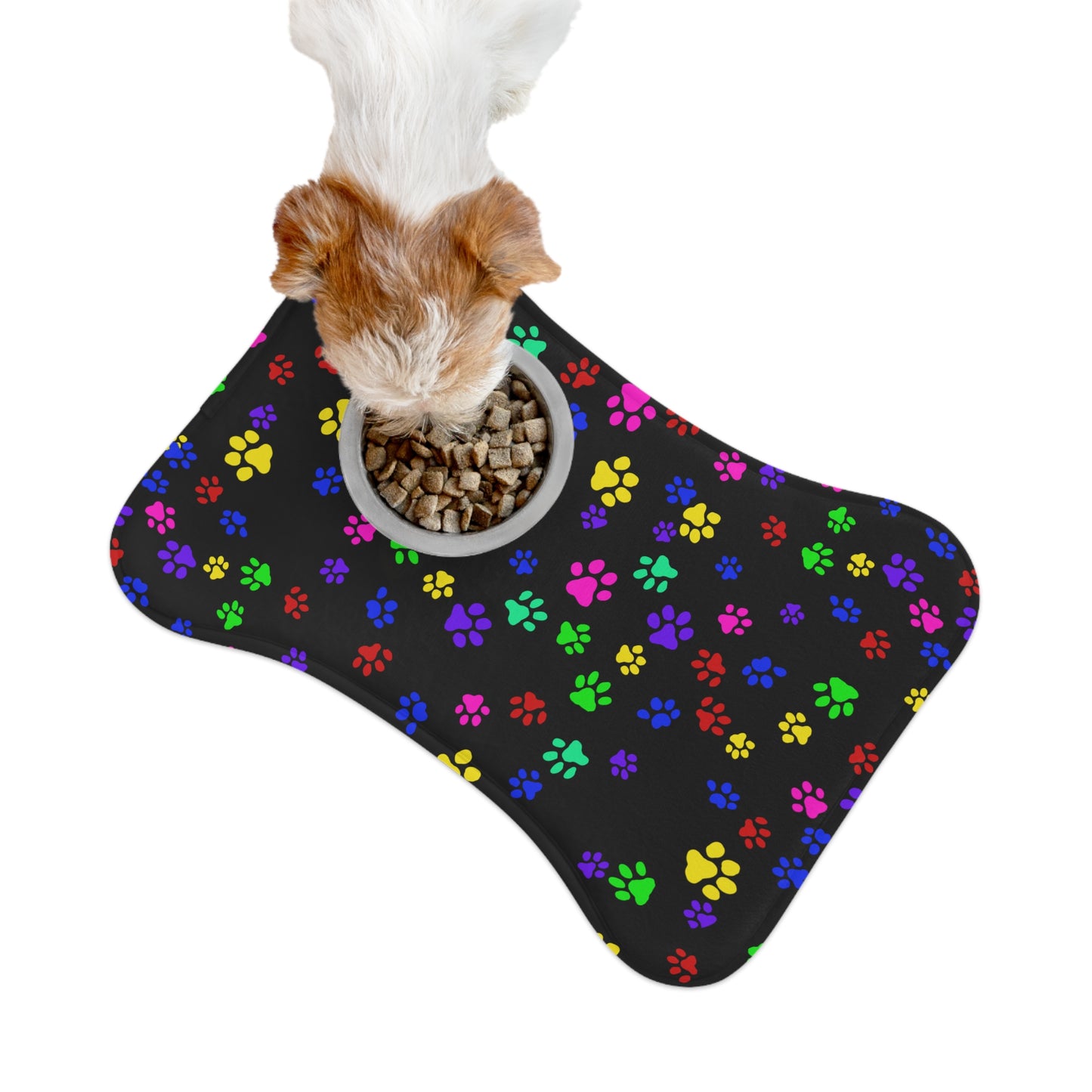 This is the 19" x 14" Bone Shape Paws on Paws Pet Feeding Mat. This shows the bone shape with different sized color paws on the design. The colors are yellow, green red, blue and purple on a black background. There is a white ceramic bowl of food on top of the right corner. The mat is around a white background and you can see the overhead view. A tan and white dog is eating out of the bowl.