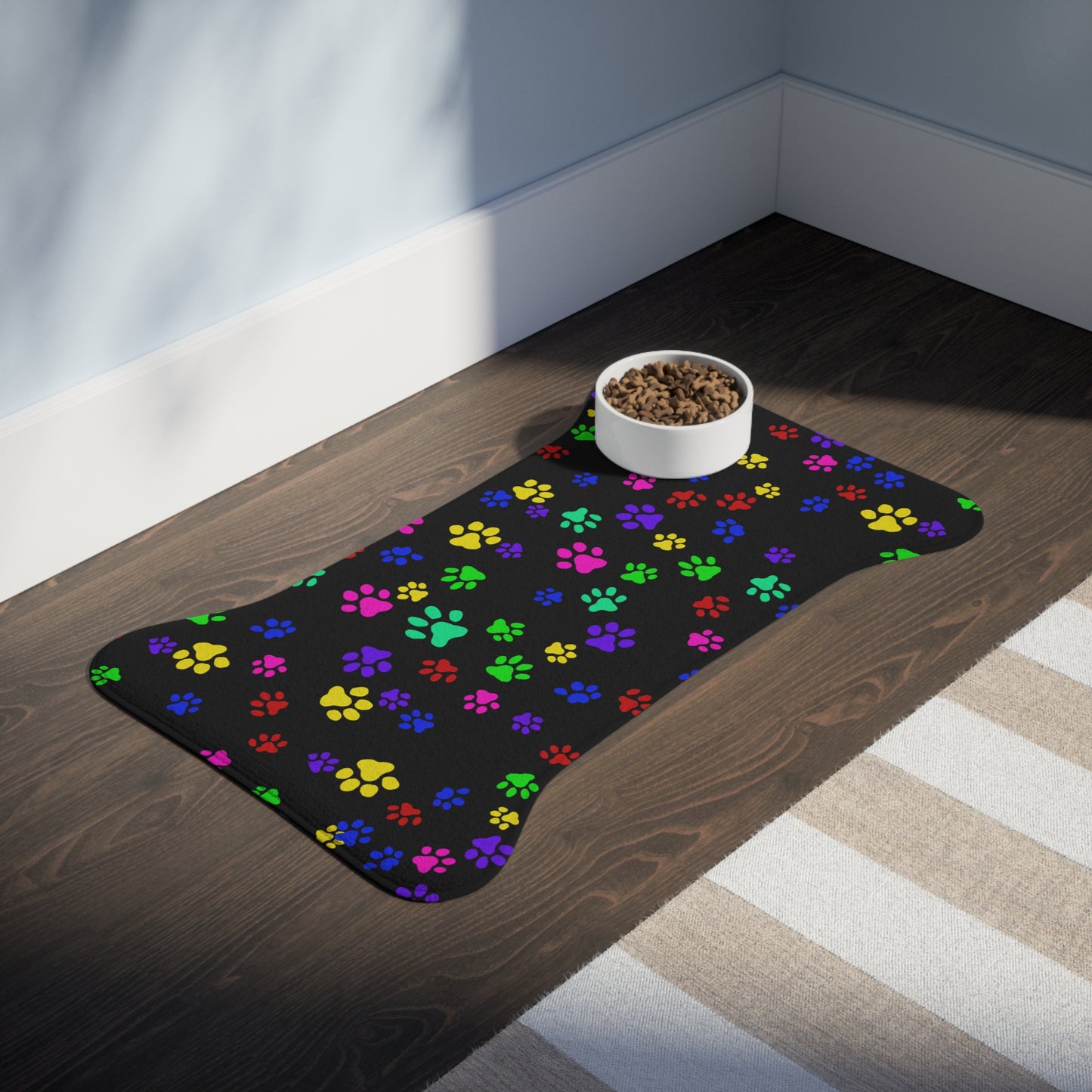 This is the 30" x 18" Bone Shape Paws on Paws Pet Feeding Mat. This shows the bone shape with different sized color paws on the design. The colors are yellow, green red, blue and purple on a black background. There is a white ceramic bowl of food on top of the right corner. The mat is placed on a hard wood floor and right below is a white and tan striped rug. The wall is a light blue and the baseboard is white.