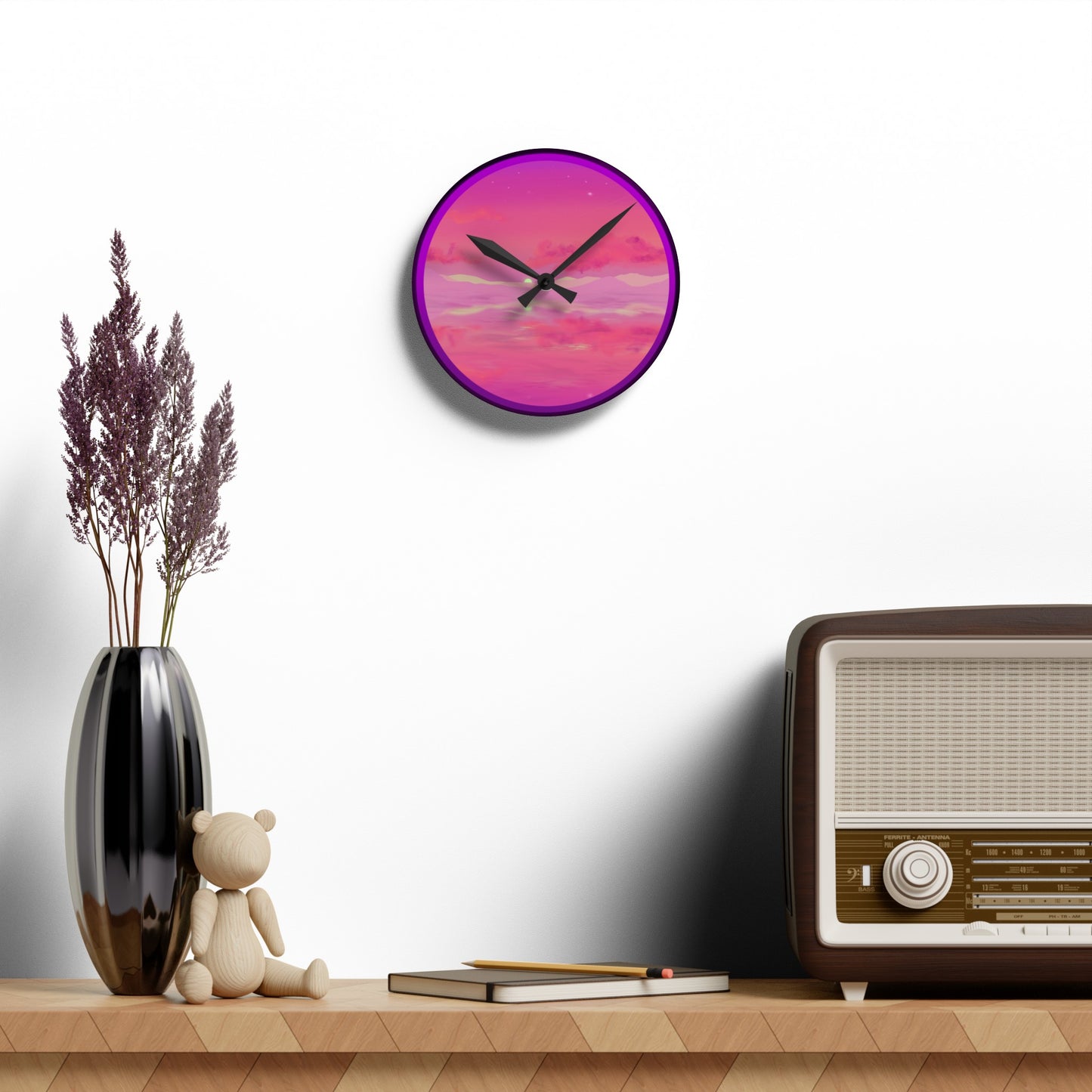 This is the 8" x 8" Pink Dremscape Acrylic Round Wall Clock by Lee Hansheng Studios.This is the front of the image with two black clock hands and the illustration behind it.The illustration has the sky and clouds. Different pinks and hues blended into the clouds and spreads outwards.There are dark purples on the side and yellows near the middle of the art piece.It is against a white background on a light wood side table with a dried plant in a shiny black oval potter,tan wood teddy bear,a journal and radio.