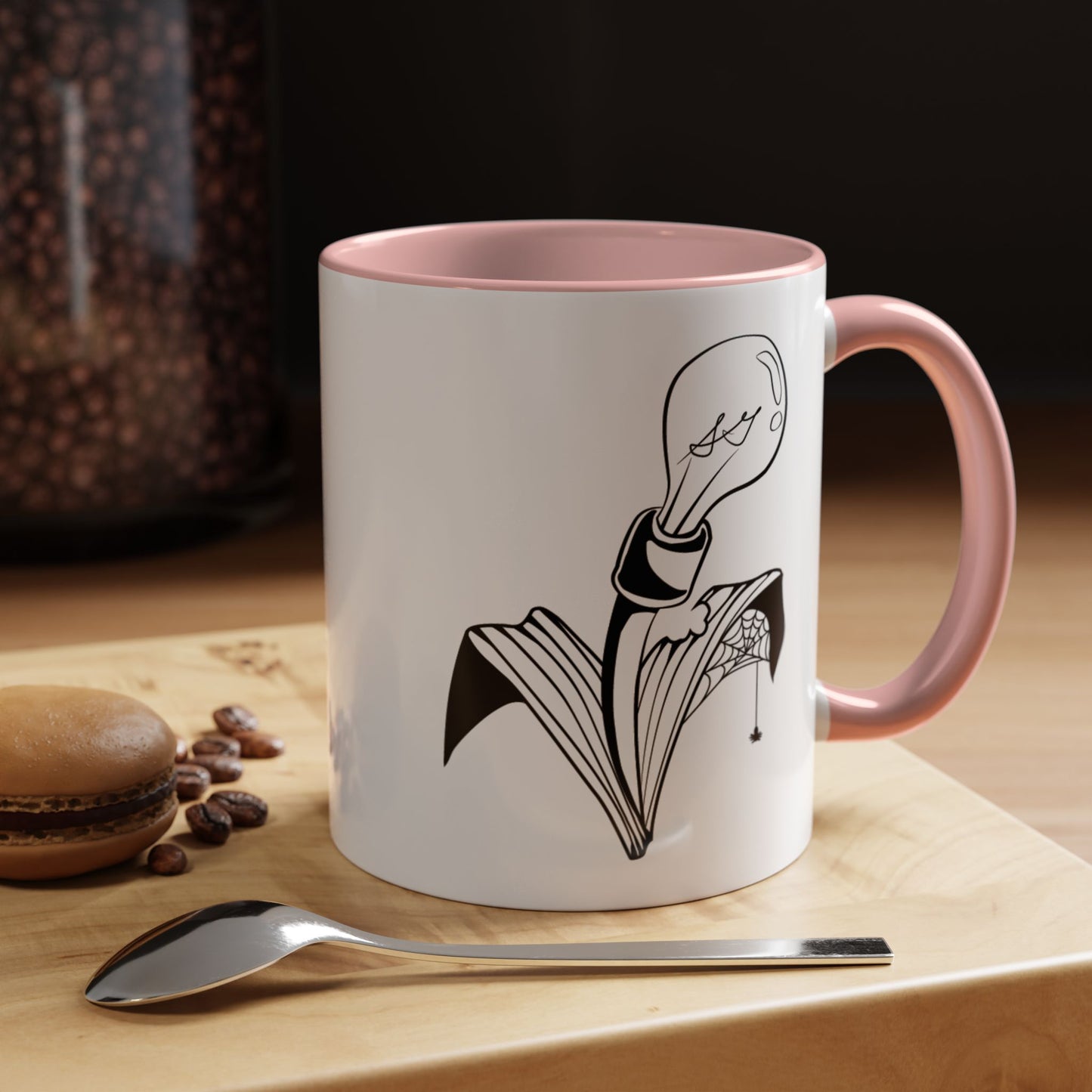 This is the Pink 11oz Storm Garden Studio Accent Coffee Mug. This is a mug by Storm Garden Studio which has the logo printed in black and white. The logo is a lightbulb that is screwed onto the leaves of a plant. One leaf is bitten off with a spiderweb. The mug is on top of a light wood block that is on top of a wood desk, a small silver spoon in the front, a chocolate macaroon and some coffee beans to the left. In the background there is a large jar of beans to the left. The wallpaper is black/grey.