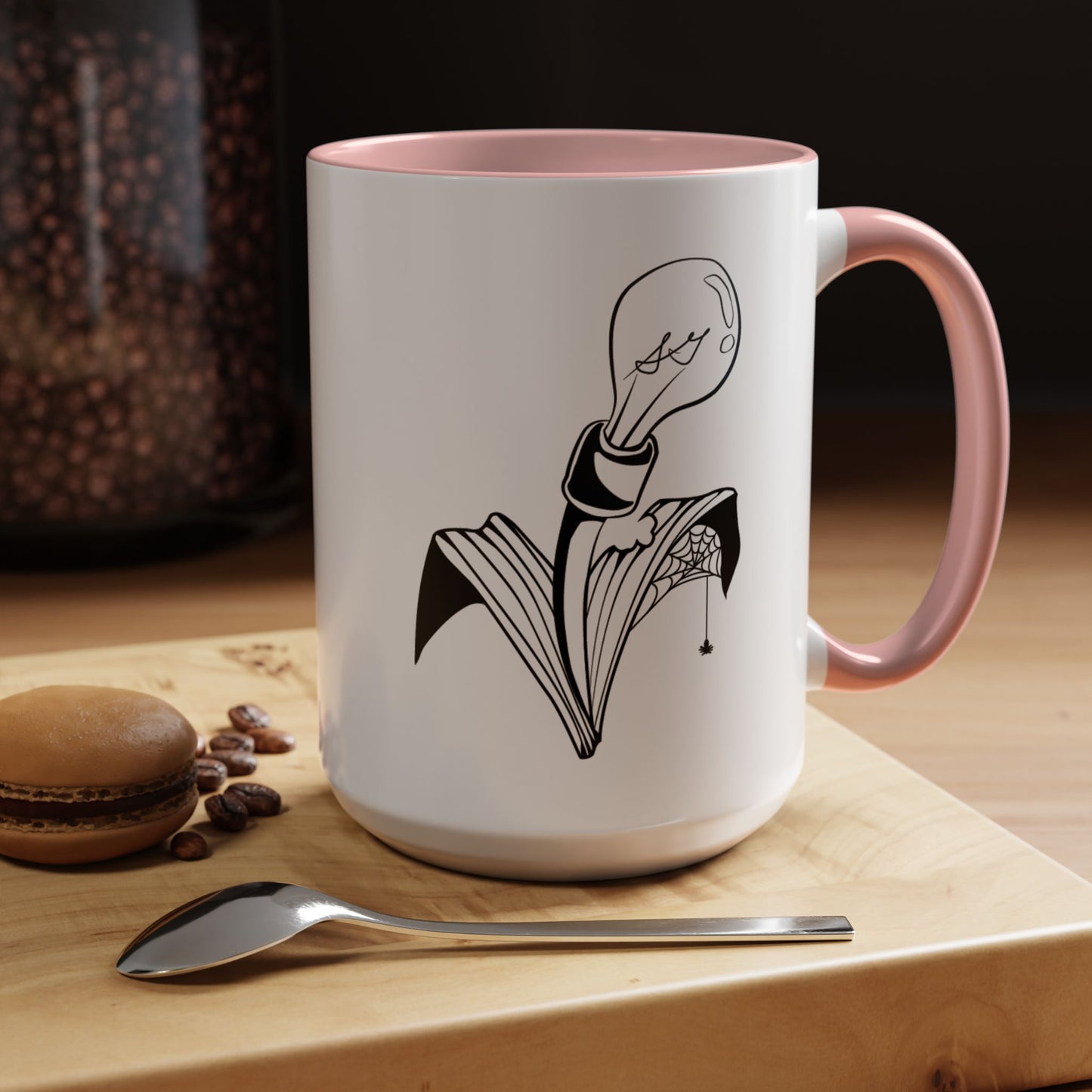 This is the Pink 15oz Storm Garden Studio Accent Coffee Mug. This is a mug by Storm Garden Studio which has the logo printed in black and white. The logo is a lightbulb that is screwed onto the leaves of a plant. One leaf is bitten off with a spiderweb. The mug is on top of a light wood block that is on top of a wood desk, a small silver spoon in the front, a chocolate macaroon and some coffee beans to the left. In the background there is a large jar of beans to the left. The wallpaper is black/grey.
