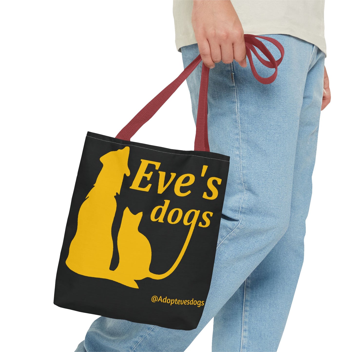 This is the Red 13" x 13" Adopt Eve's Dog Tote Bag. The handles are red. The top of the tote is hemmed in yellow. The tote is all over black with the image of a dog, cat, "Eve's Dogs" and "@Adoptevesdogs" in yellow. A man's hand with a beige tee and light- colored jeans is grabbing the handles from the hip down. The tote is against a white background.