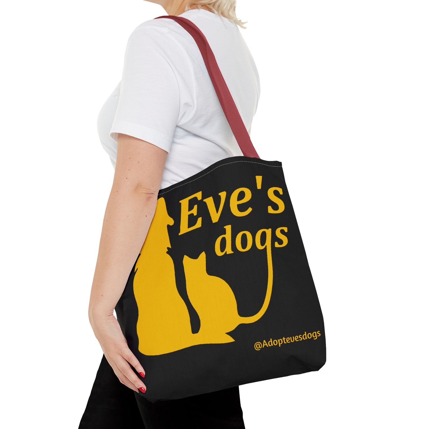 This is the Red 16" x 16" Adopt Eve's Dog Tote Bag. The handles are red. The top of the tote is hemmed in yellow. The tote is all over black with the image of a dog, cat, "Eve's Dogs" and "@Adoptevesdogs" in yellow. It is over the shoulder of a short blonde haired woman wearing a white tea and black pants. Her hand with red nails is on the tote and she is facing the left.