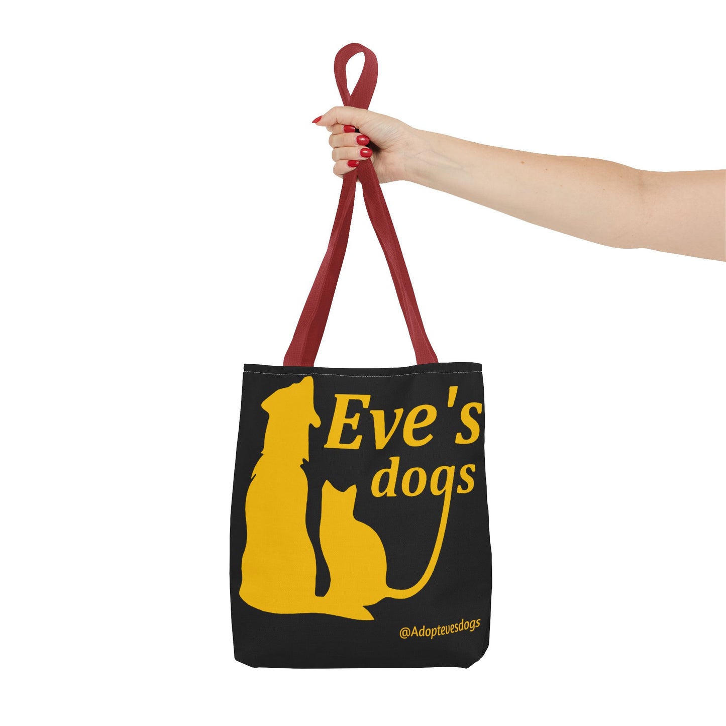 This is the Red 16" x 16" Adopt Eve's Dog Tote Bag. The handles are red. The top of the tote is hemmed in yellow. The tote is all over black with the image of a dog, cat, "Eve's Dogs" and "@Adoptevesdogs" in yellow. A woman's hand with red nail polish is grabbing the handles. Her arm is outstretched to the middle. The tote is against a white background.