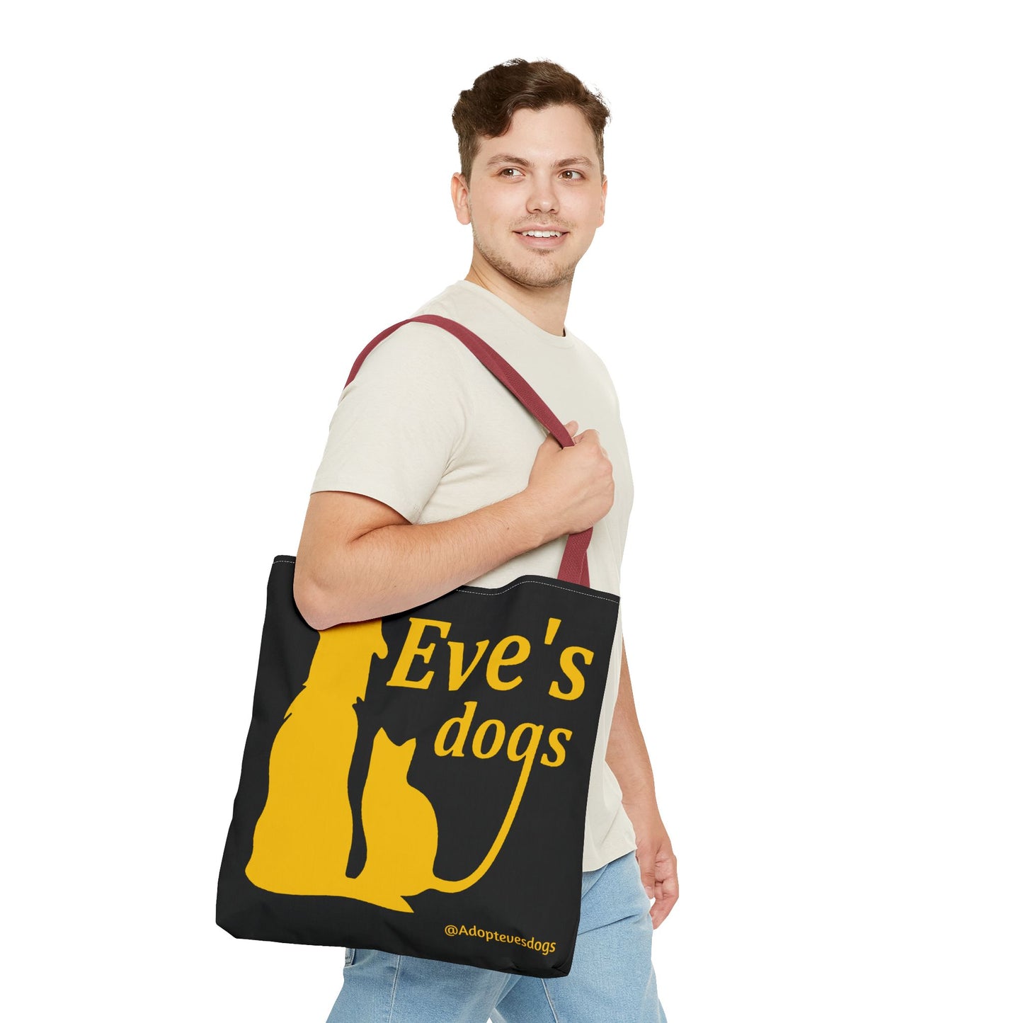 This is the Red 16" x 16" Adopt Eve's Dog Tote Bag. The handles are red. The top of the tote is hemmed in yellow. The tote is all over black with the image of a dog, cat, "Eve's Dogs" and "@Adoptevesdogs" in yellow. A man with a smile, short wavy brown hair, beige tee plus jeans with tote around their shoulder and walking to the right against a white background.