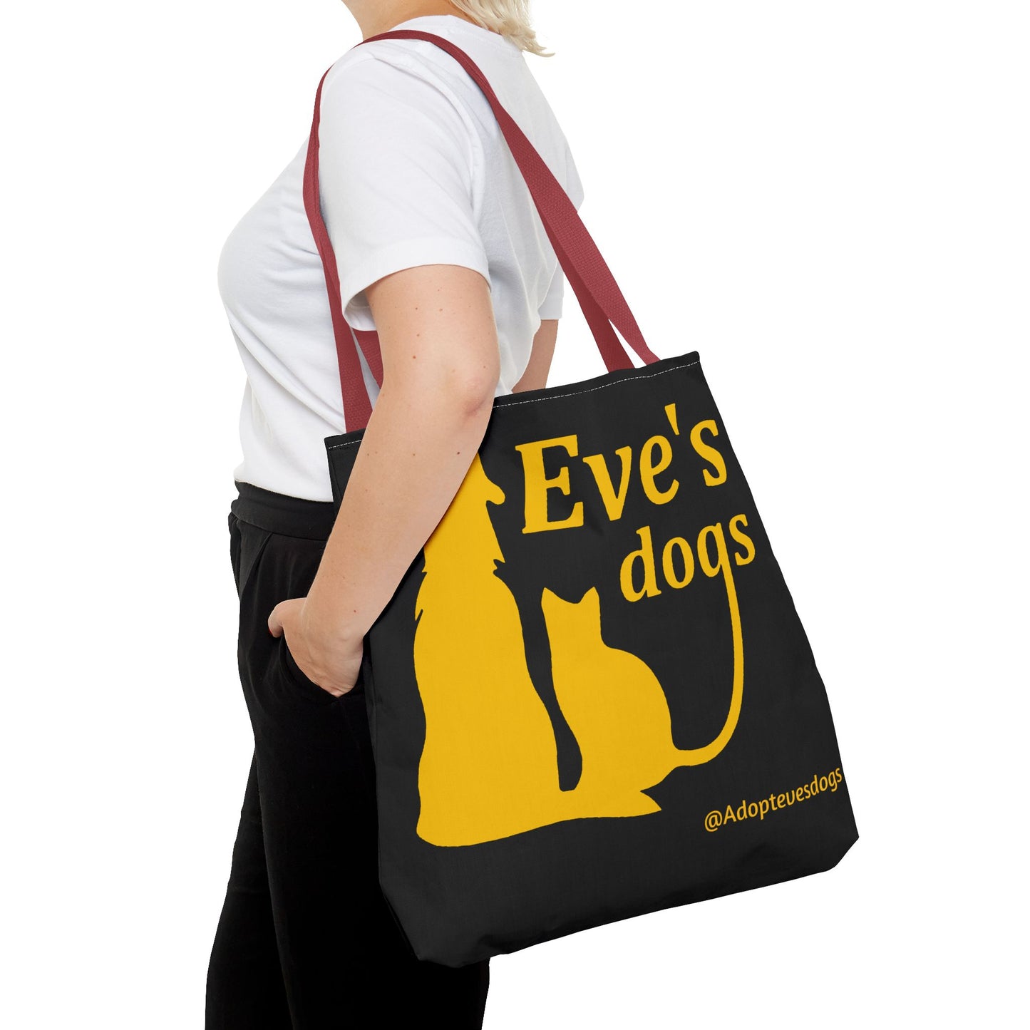 This is the Red 18" x 18" Adopt Eve's Dog Tote Bag. The handles are red. The top of the tote is hemmed in yellow. The tote is all over black with the image of a dog, cat, "Eve's Dogs" and "@Adoptevesdogs" in yellow. It is over the shoulder of a short blonde haired woman wearing a white tea and black pants. Her hand is in the pant's pocket and she is facing the left.