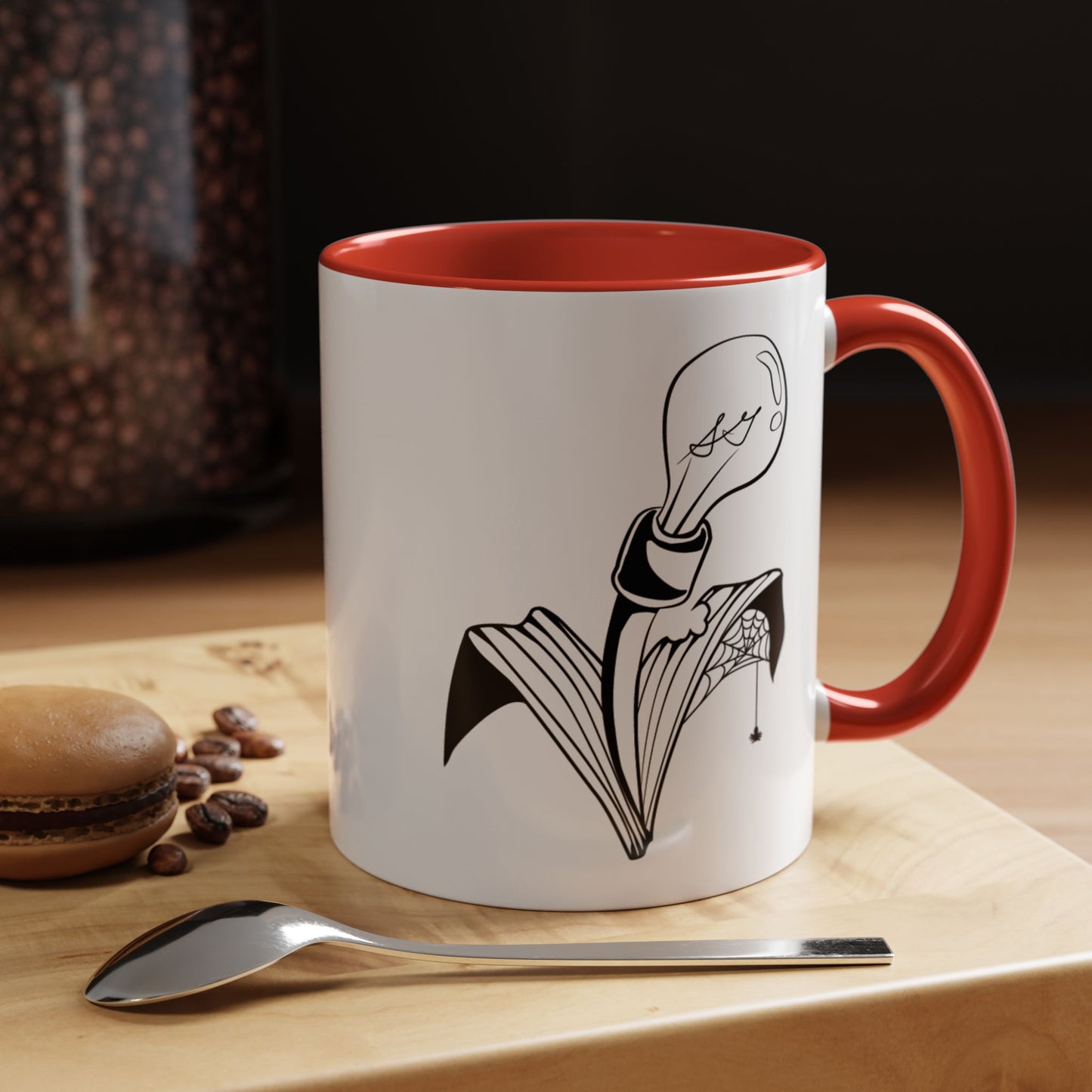 This is the Red 11oz Storm Garden Studio Accent Coffee Mug. This is a mug by Storm Garden Studio which has the logo printed in black and white. The logo is a lightbulb that is screwed onto the leaves of a plant. One leaf is bitten off with a spiderweb. The mug is on top of a light wood block that is on top of a wood desk, a small silver spoon in the front, a chocolate macaroon and some coffee beans to the left. In the background there is a large jar of beans to the left. The wallpaper is black/grey.